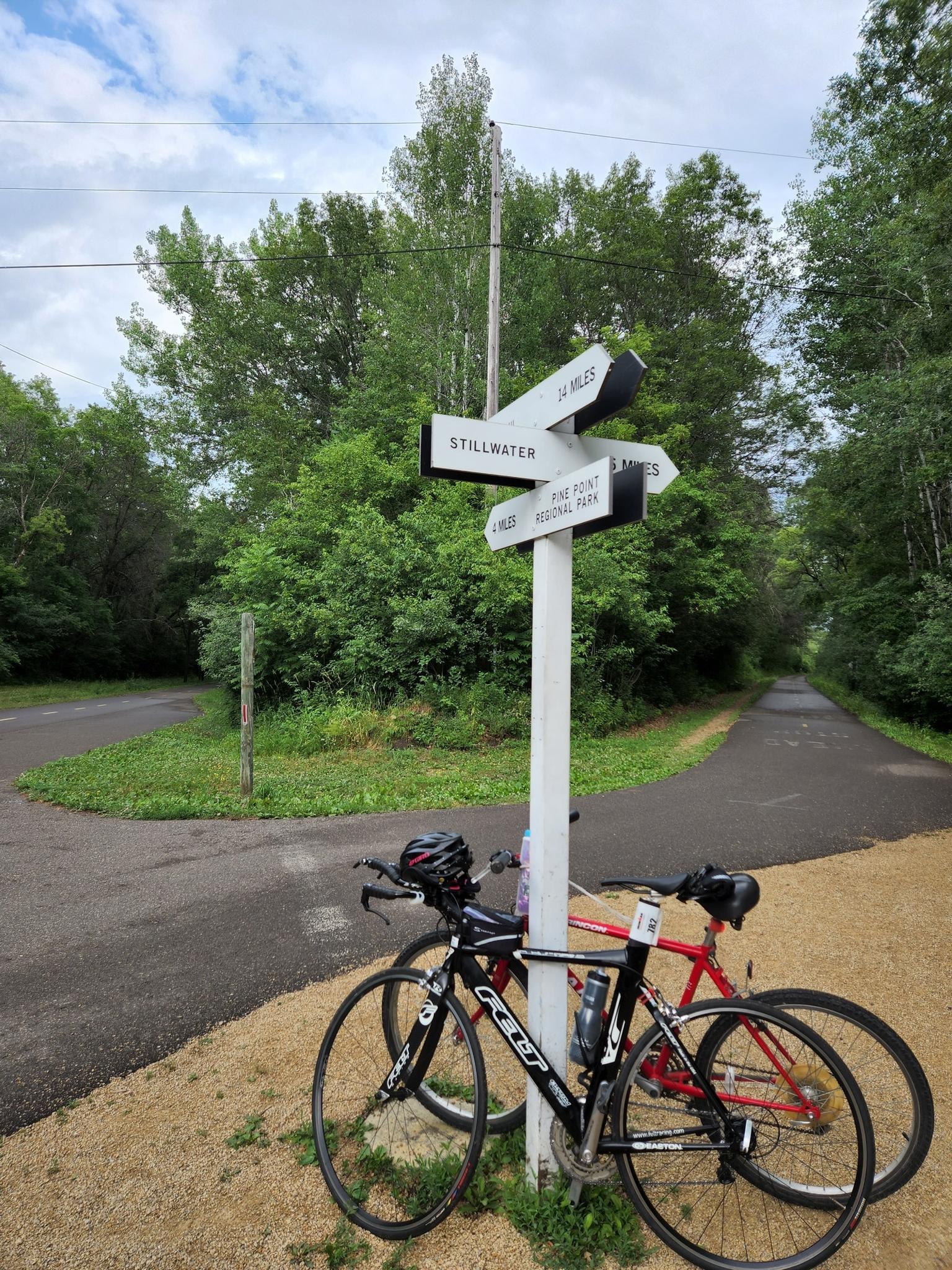 Two bicycles parked next to a signpost on a paved path in a forest.