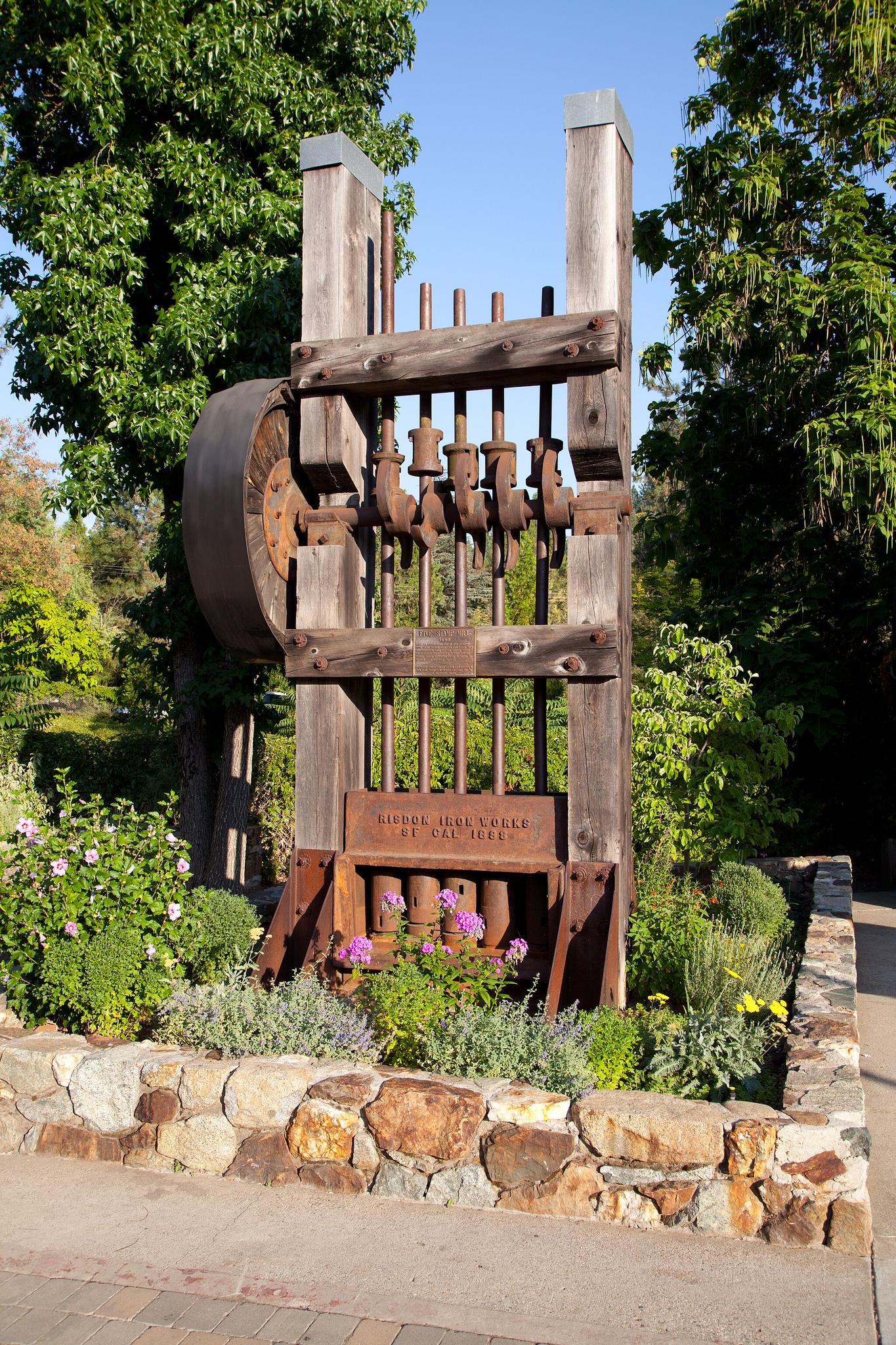 A large wooden and metal sculpture surrounded by green plants and flowers.