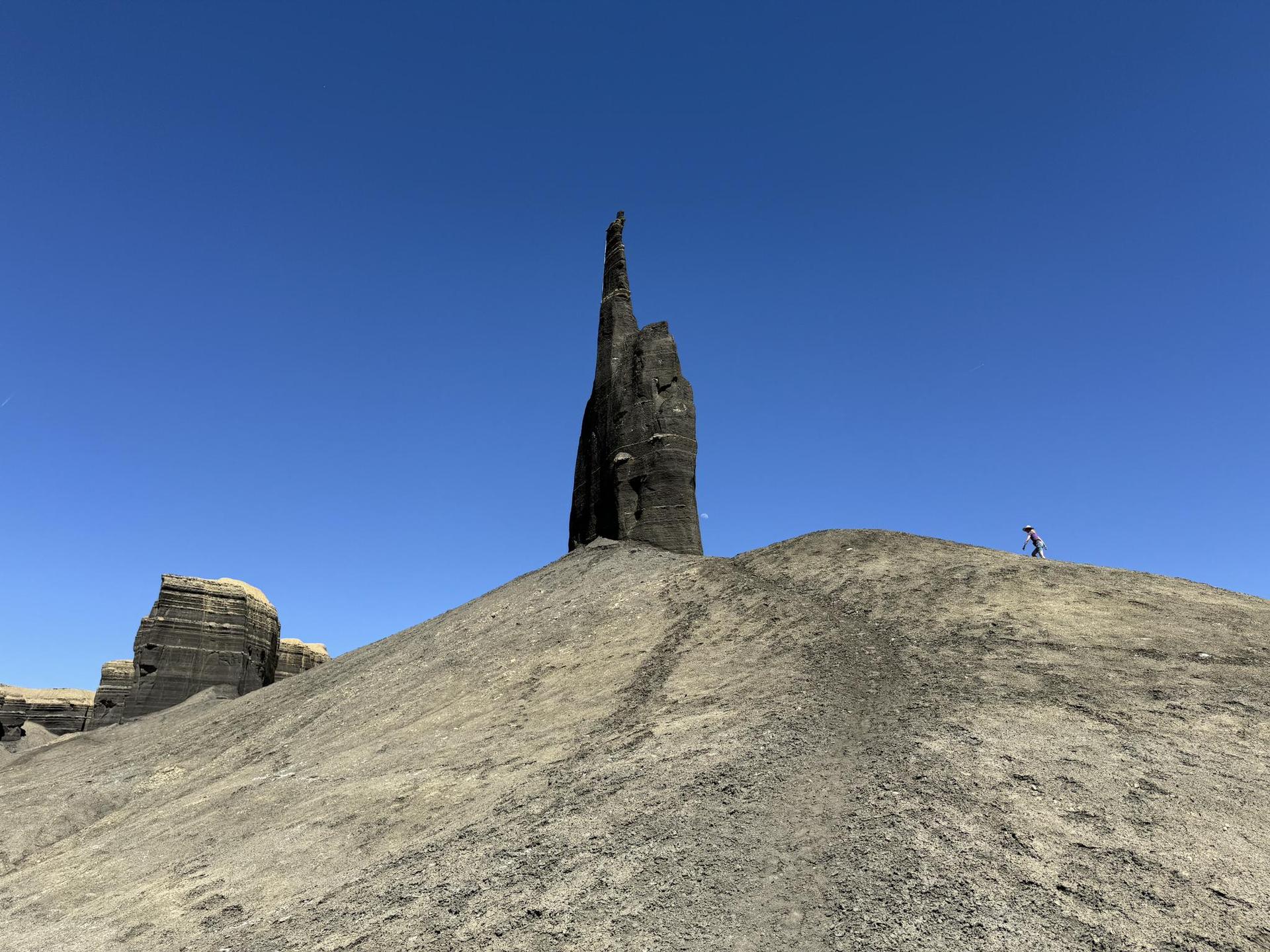 A person hikes near a tall rock spire in a sunny, arid landscape.