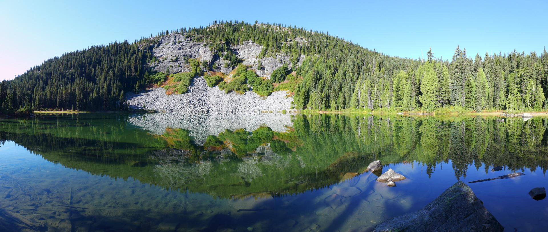 A calm lake perfectly reflects a sunlit, tree-covered mountain under a blue sky.