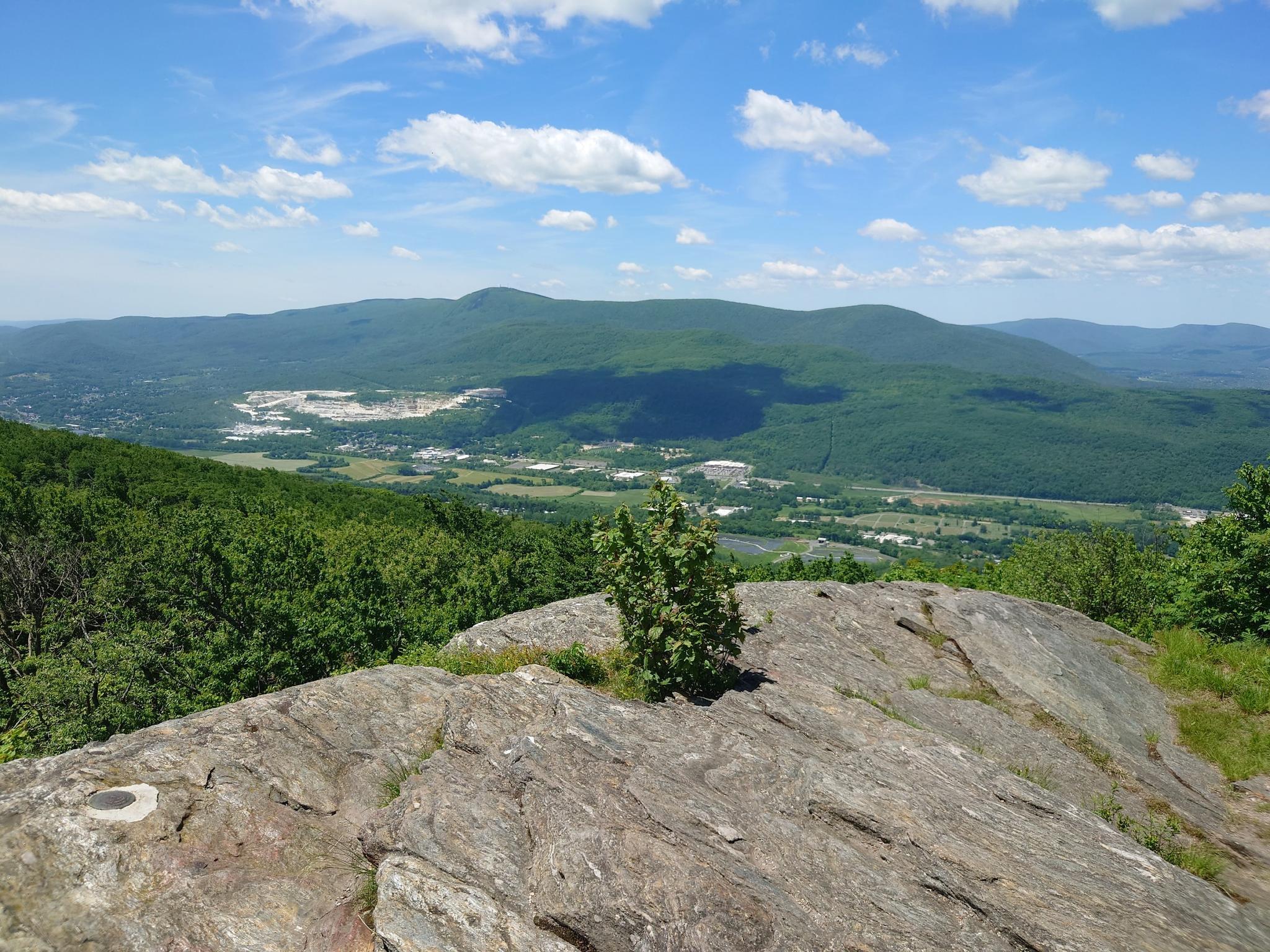 Sunny view from a rocky outcrop overlooking a forested valley and distant mountains.
