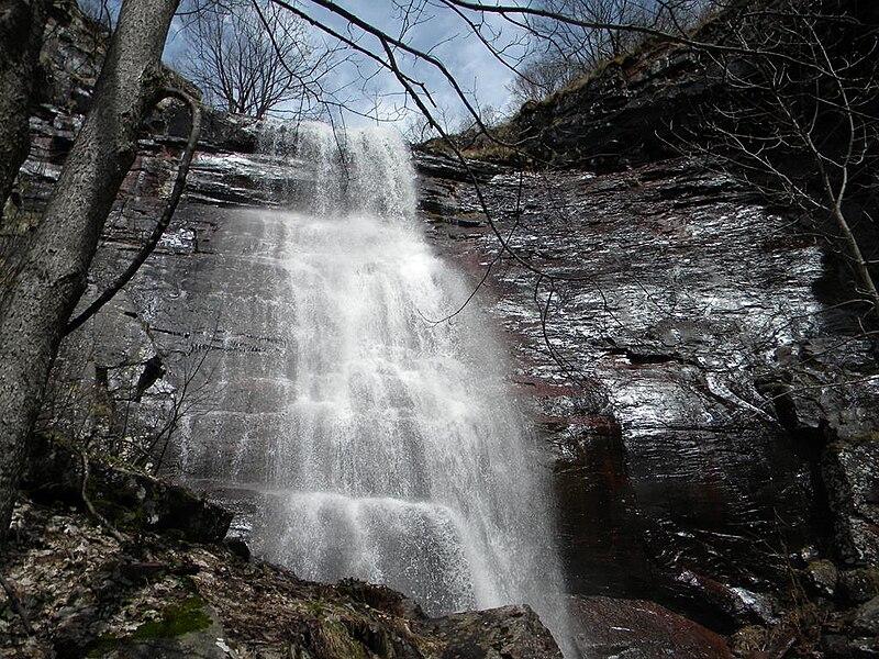 Tall waterfall cascades down dark rocky cliffs with bare trees under a bright sky.