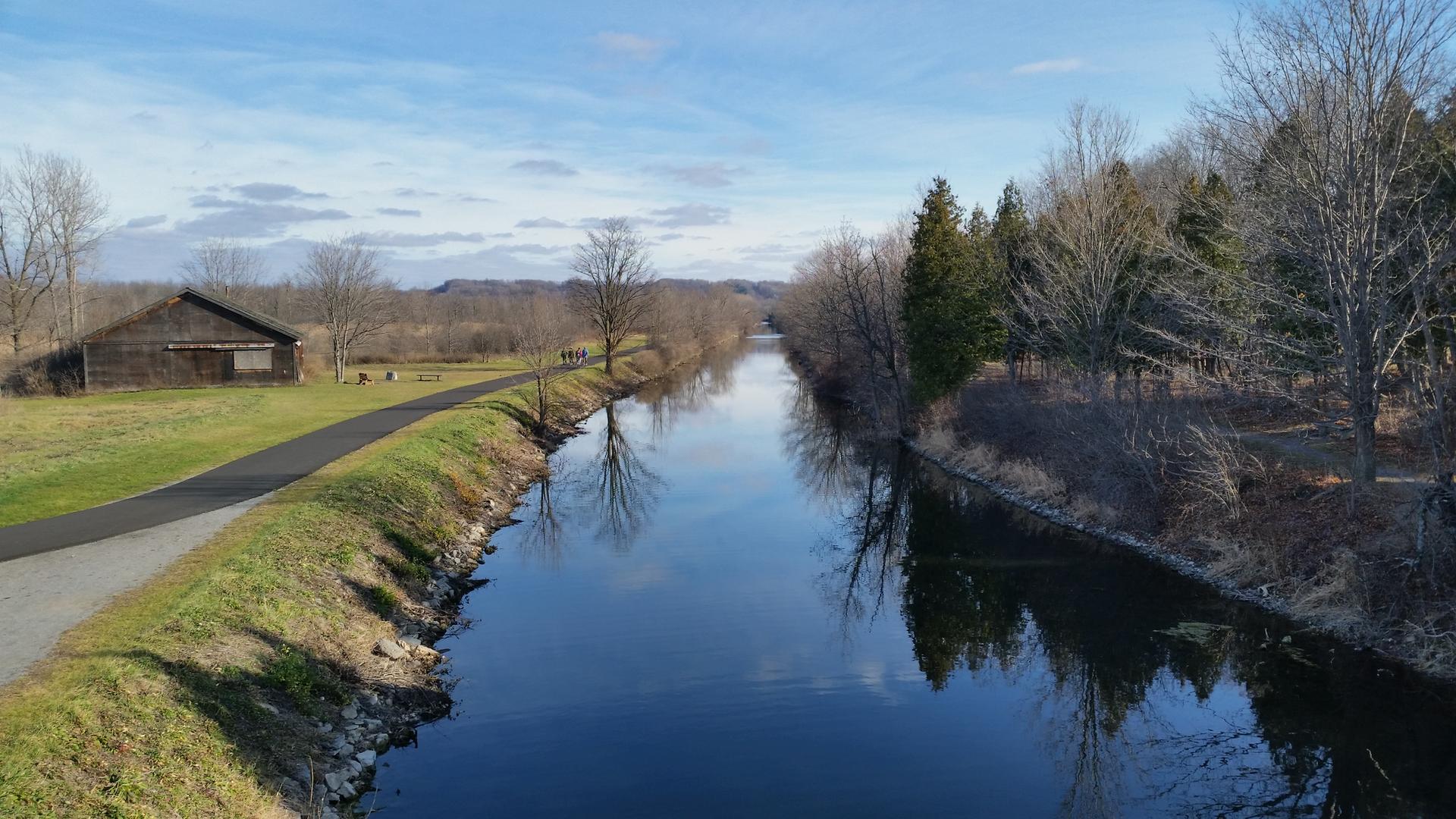 A calm canal with a path, bare trees, and a building on a clear day.