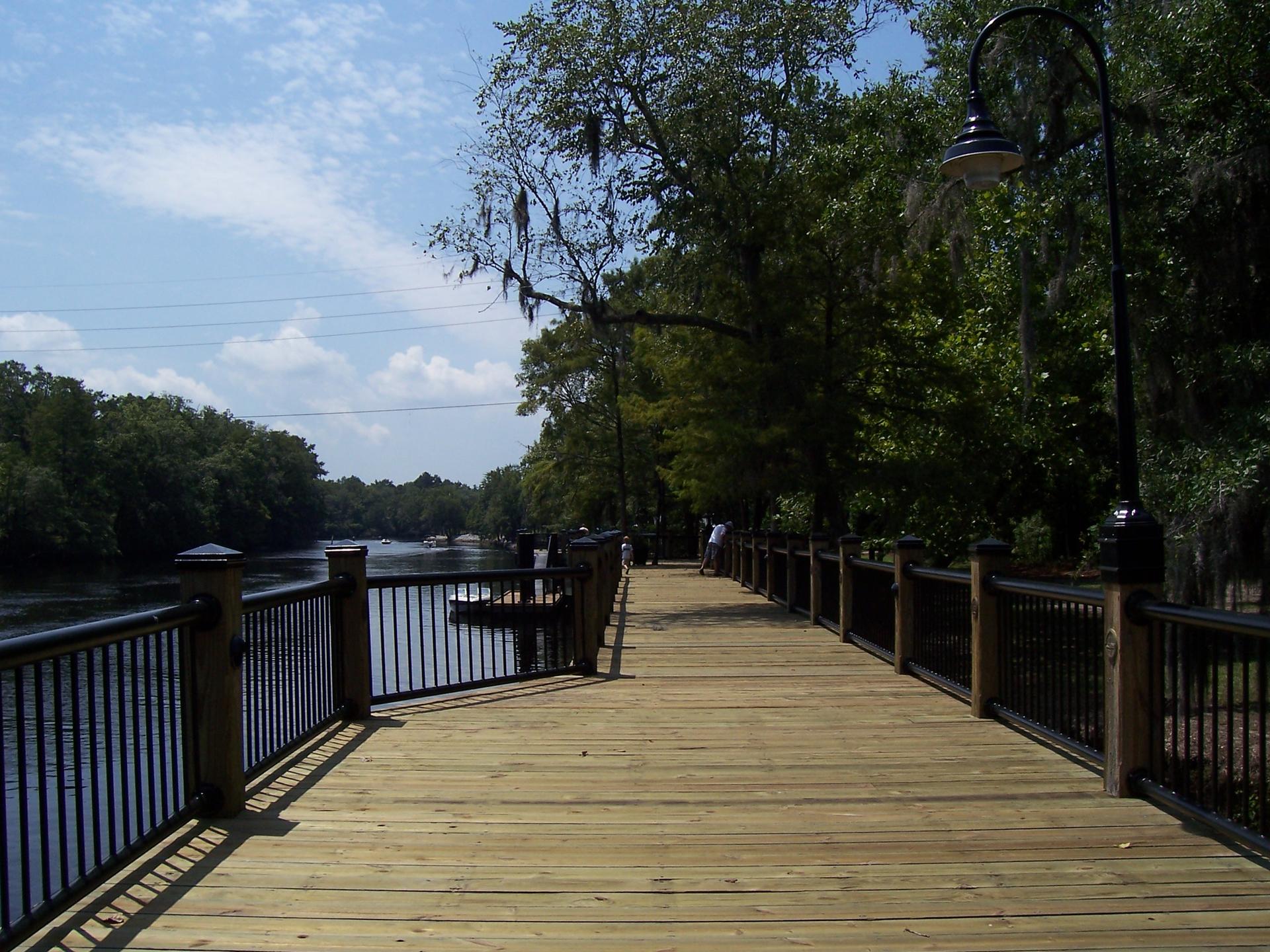 People walk on a sunny wooden boardwalk alongside a river with lush trees.