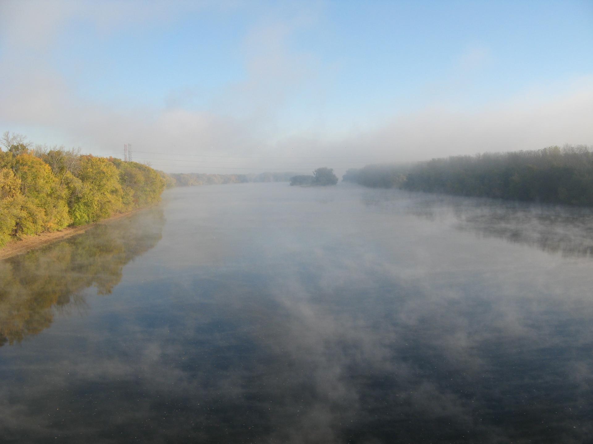 Misty river at dawn, flanked by dense green and autumn trees.