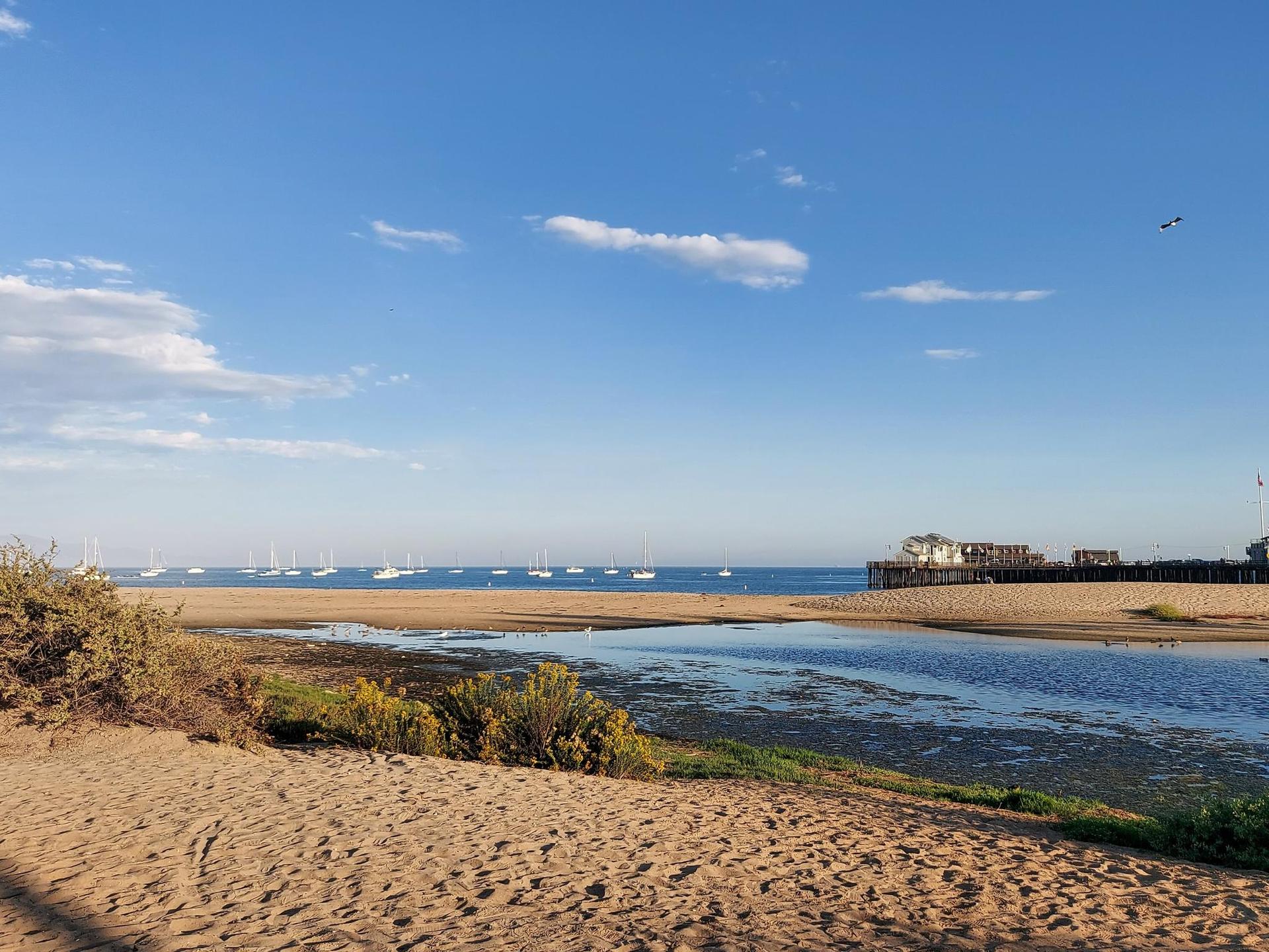 Beach with a tidal inlet, distant pier, and boats under a blue sky.