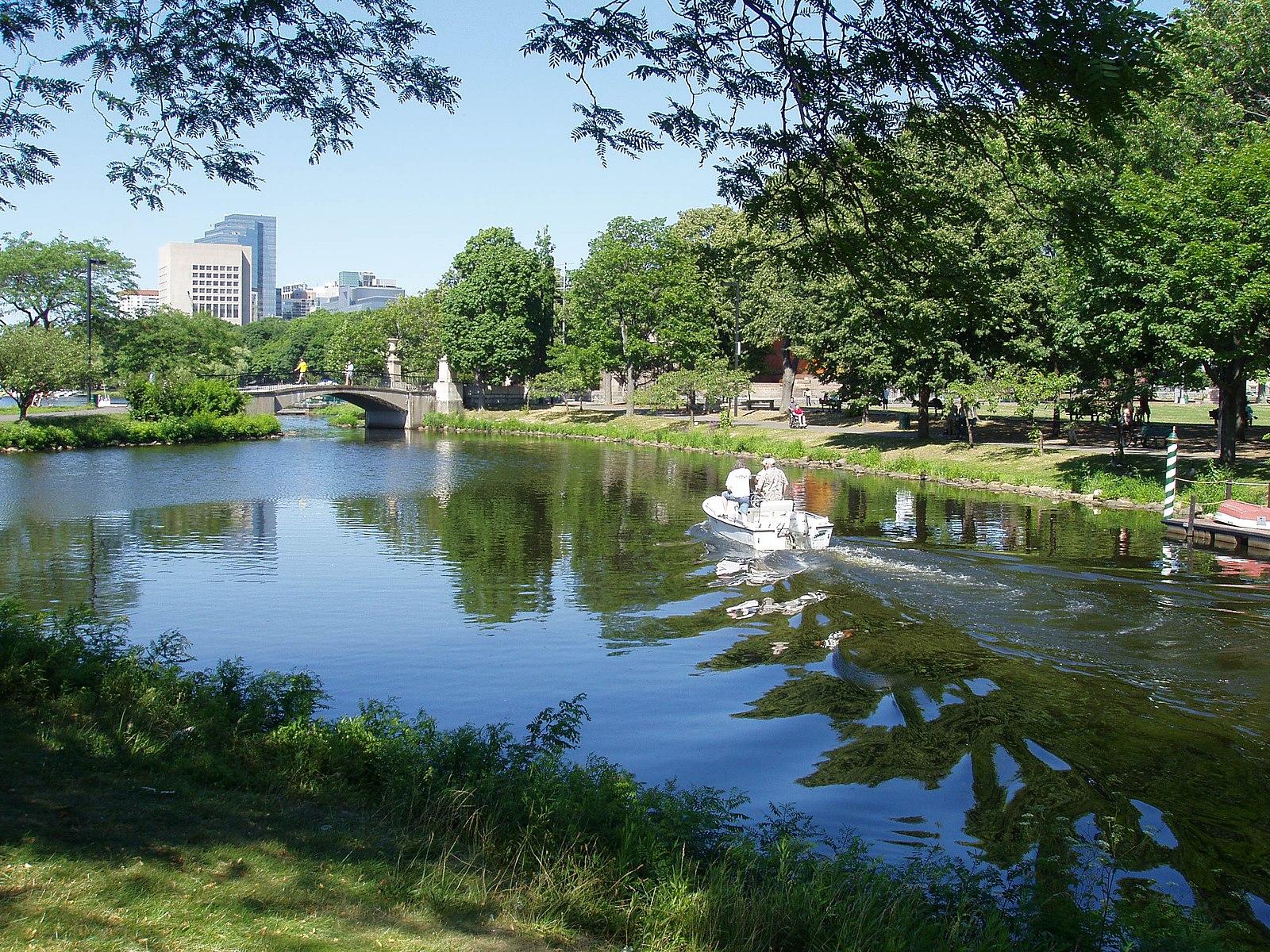 Small boat with two people on a sunny river, city buildings in background.