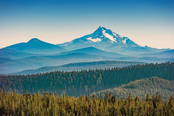 Snow-capped mountain peak rises above layers of forested hills under a clear sky.