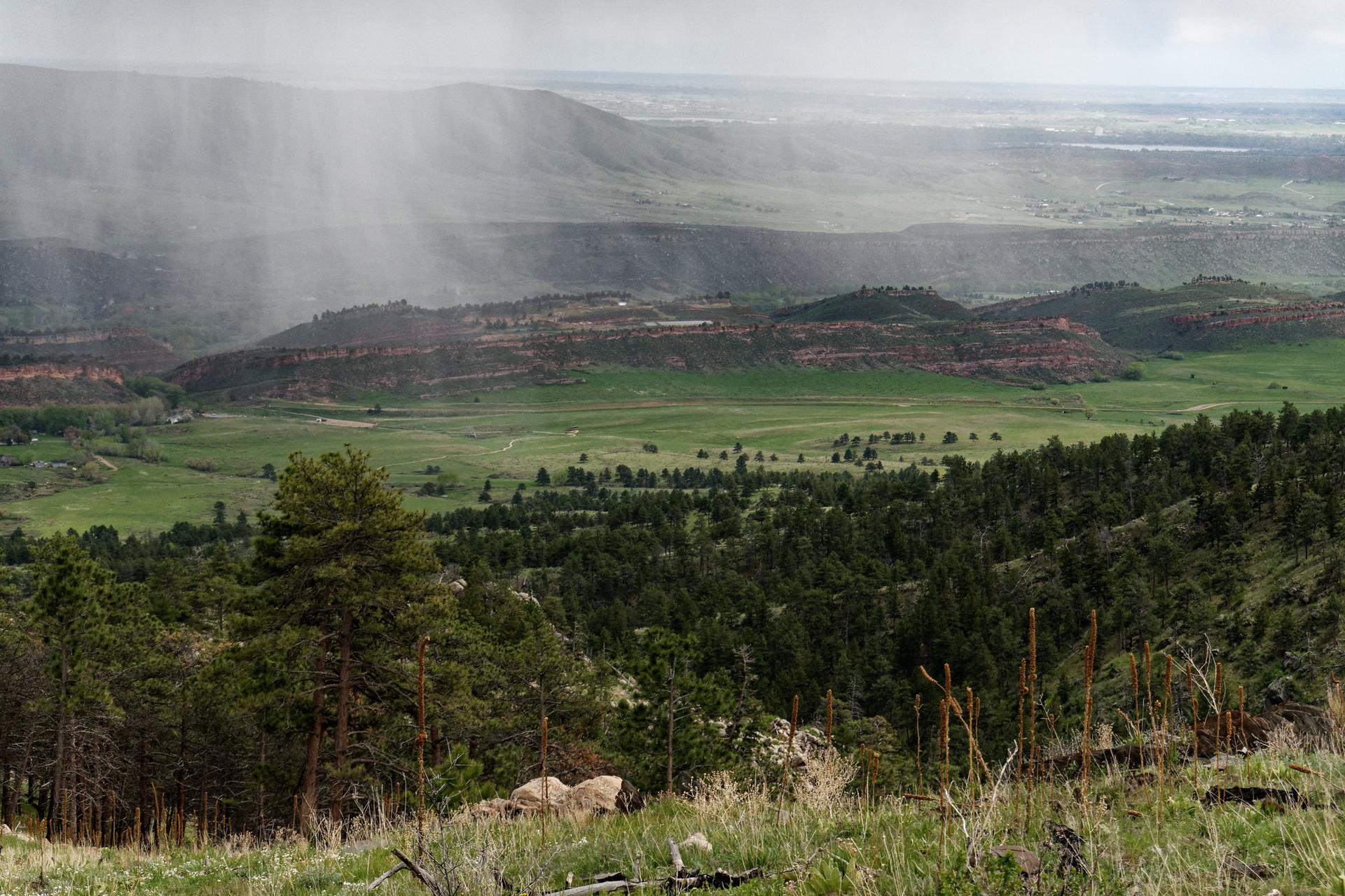 Rain shafts fall over a vast green valley with forested hills and distant mountains.