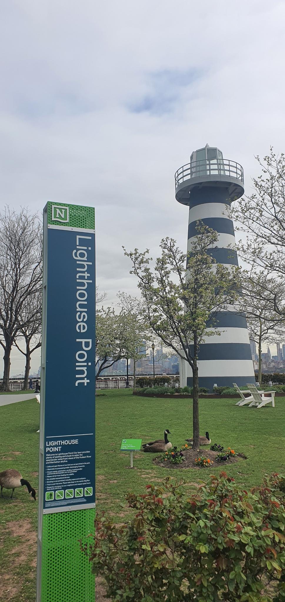 Striped lighthouse and a green sign in a park with geese on an overcast day.