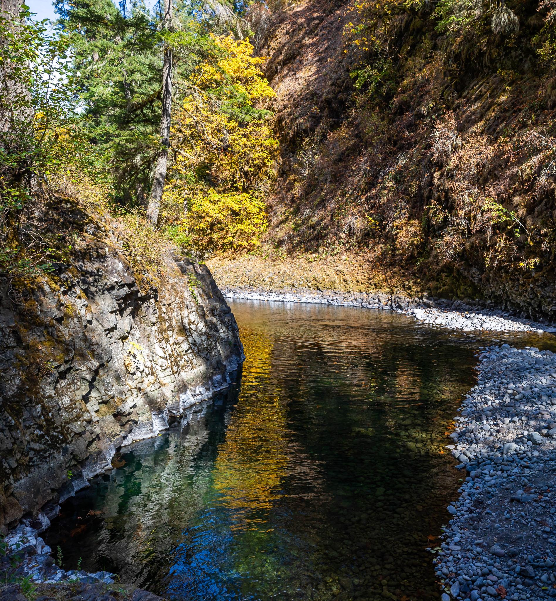 A clear river flows past rocky cliffs and vibrant autumn trees in dappled sunlight.
