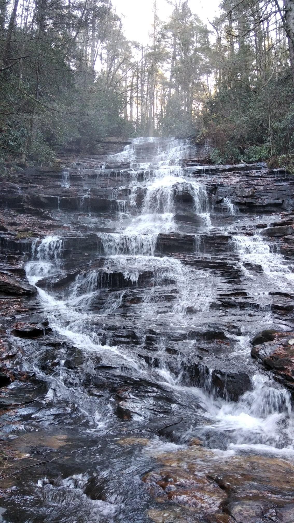 Natural light illuminates a cascading waterfall over dark, layered rock formations in a forest.