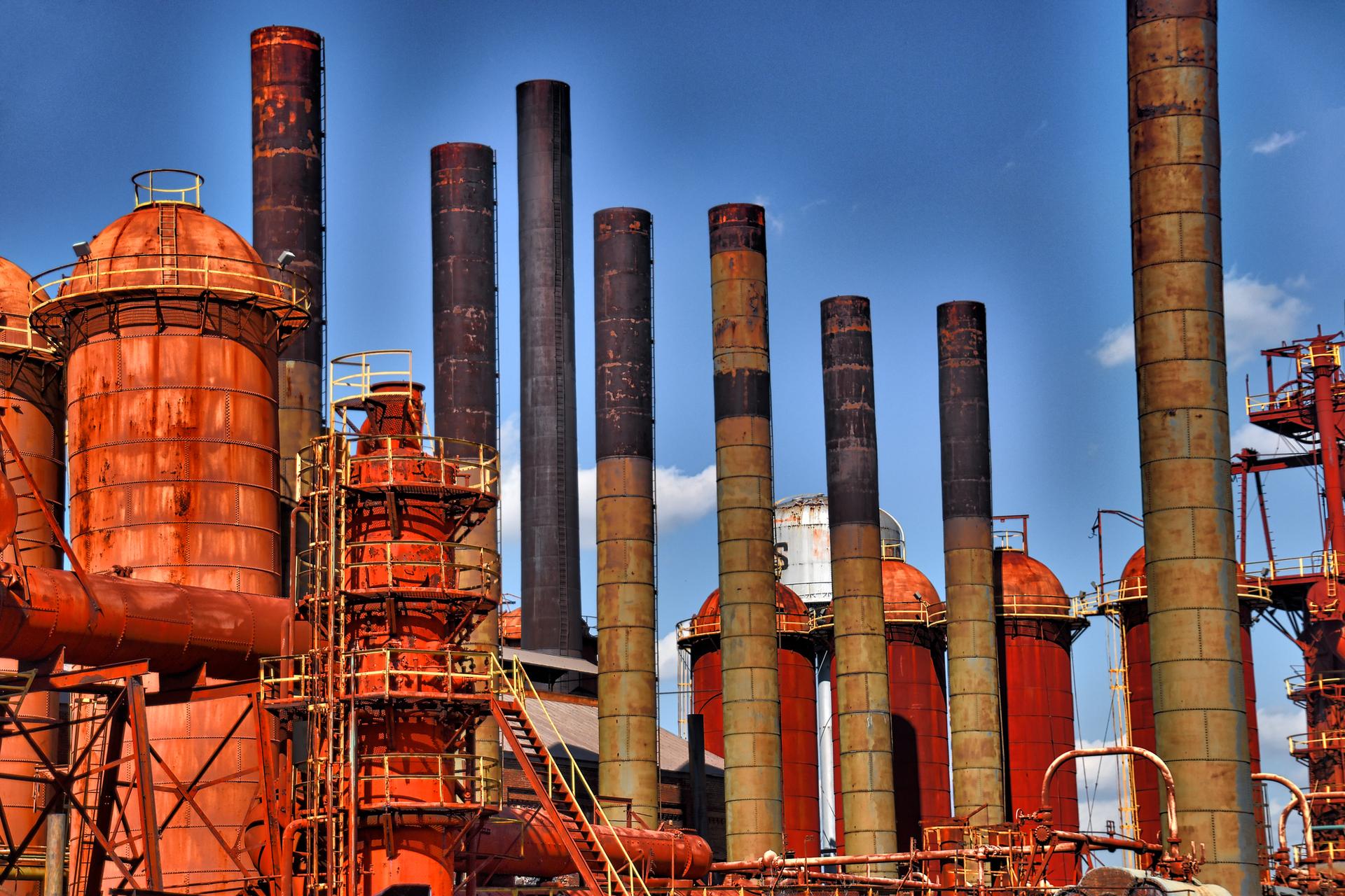 Rusty industrial complex with numerous tall smokestacks under a bright blue sky.
