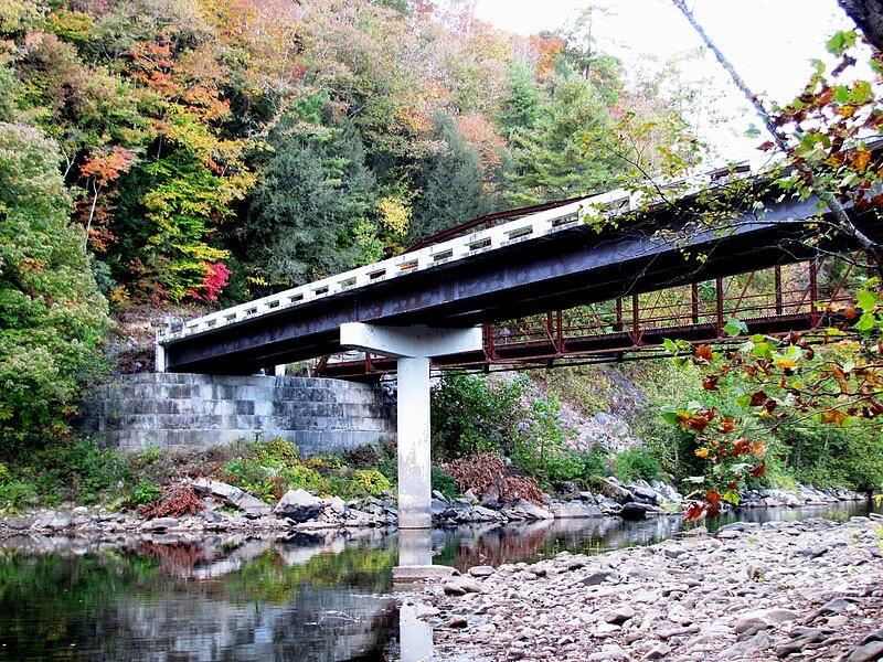 A bridge crosses a river, flanked by vibrant autumn trees under natural light.
