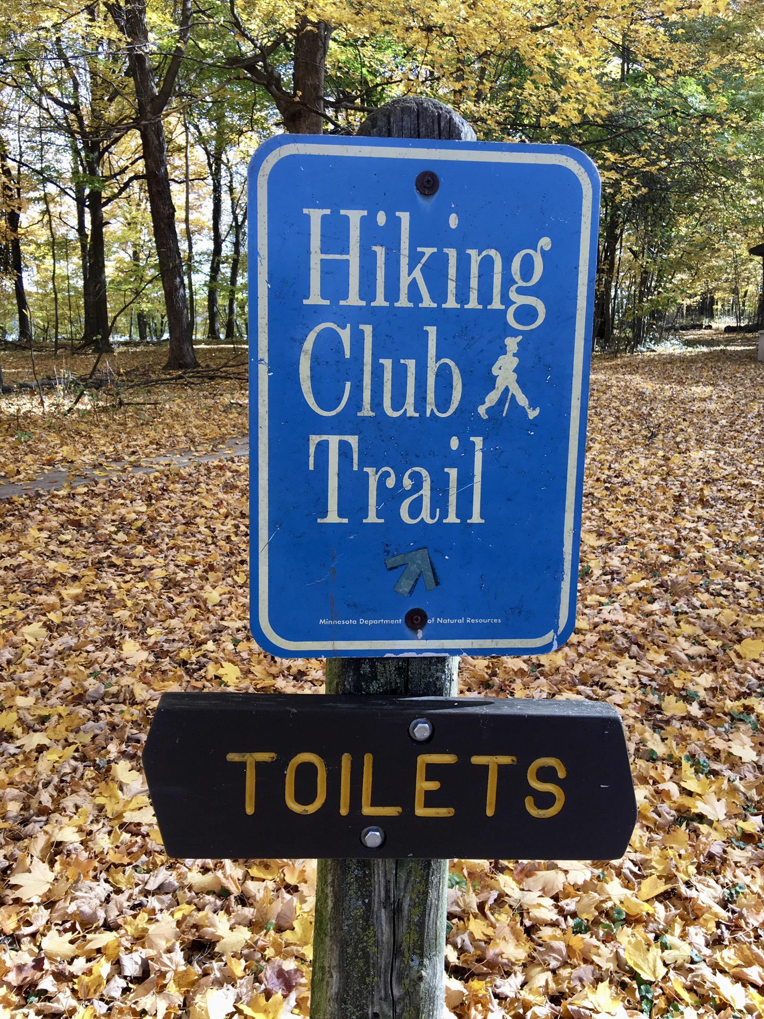 Blue hiking trail sign and black toilets sign amidst fallen autumn leaves.