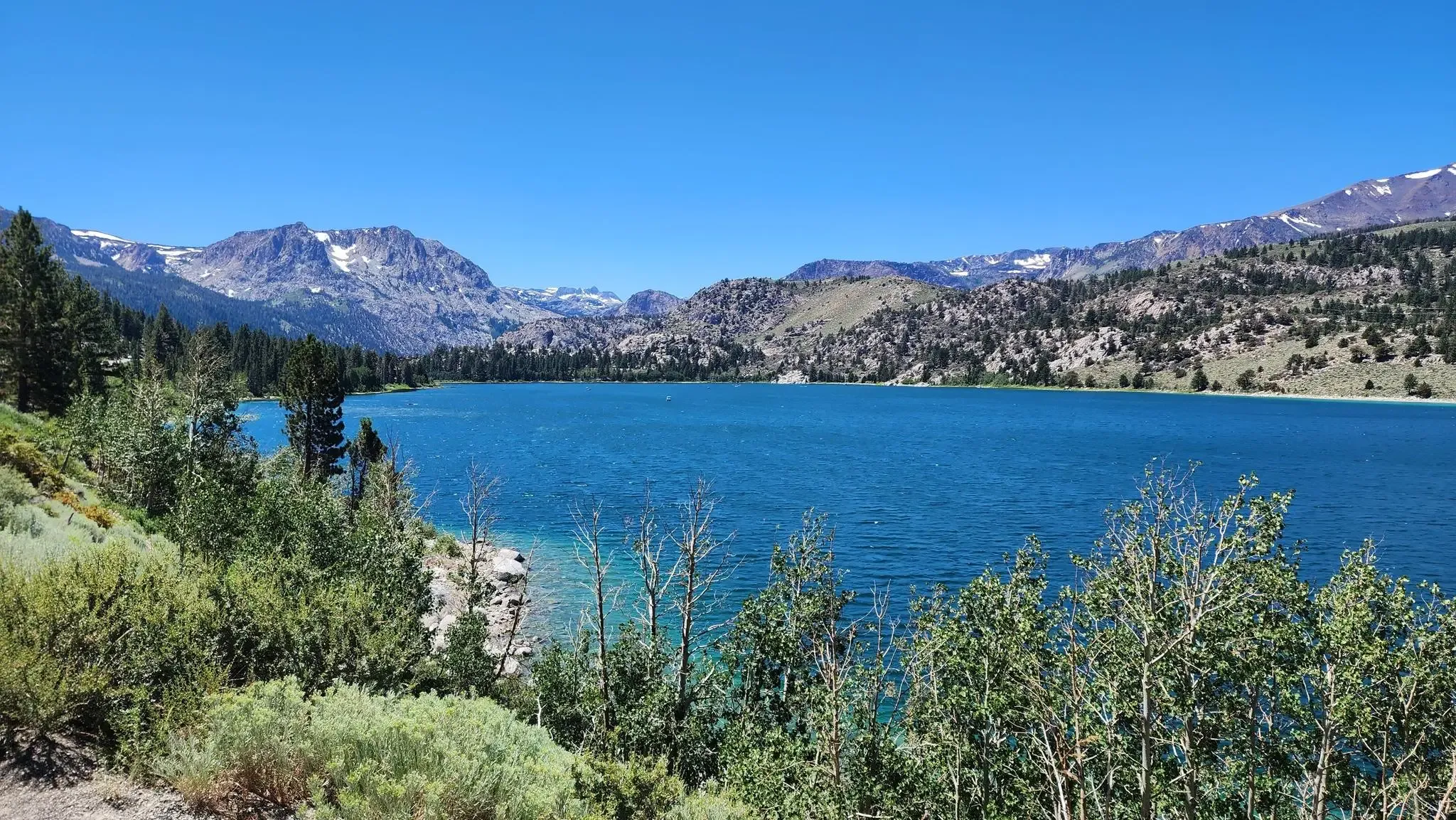 Bright blue lake with green trees and distant mountains under a clear sky. - Yost Meadow Trail