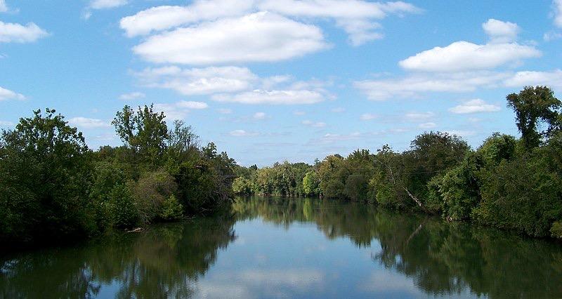 Serene river reflecting blue sky and white clouds, flanked by green trees.