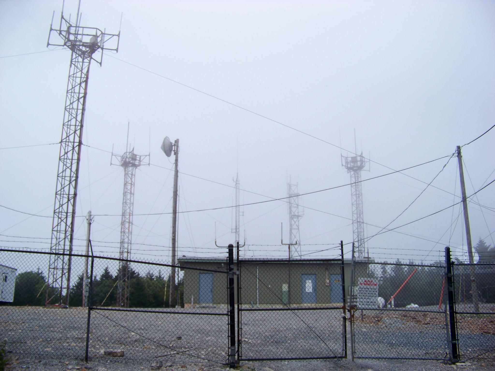Communication towers and a building in dense fog behind a chain-link fence.