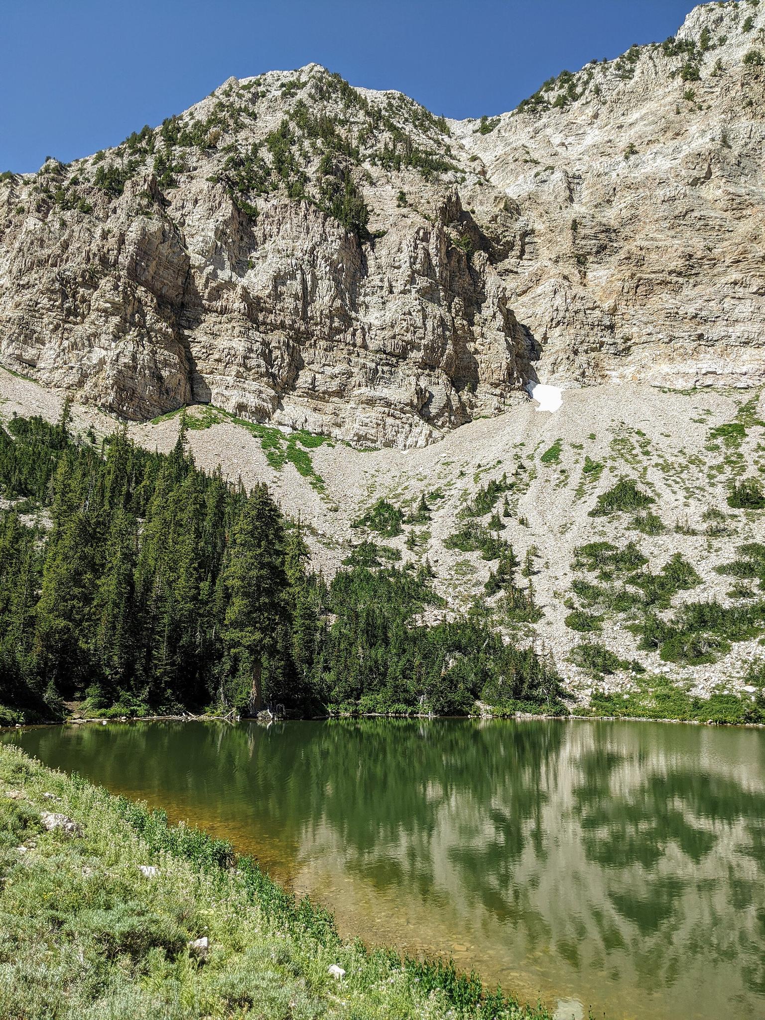Clear mountain lake reflecting rocky cliffs and pine trees on a sunny day.