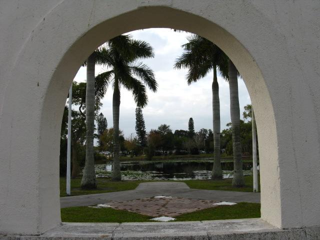 View through a white archway to palm trees, a pond, and green grass.