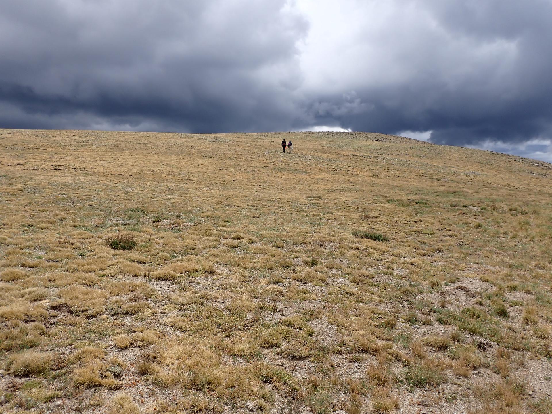 Two hikers ascend a vast, dry, grassy hill under dramatic dark clouds.