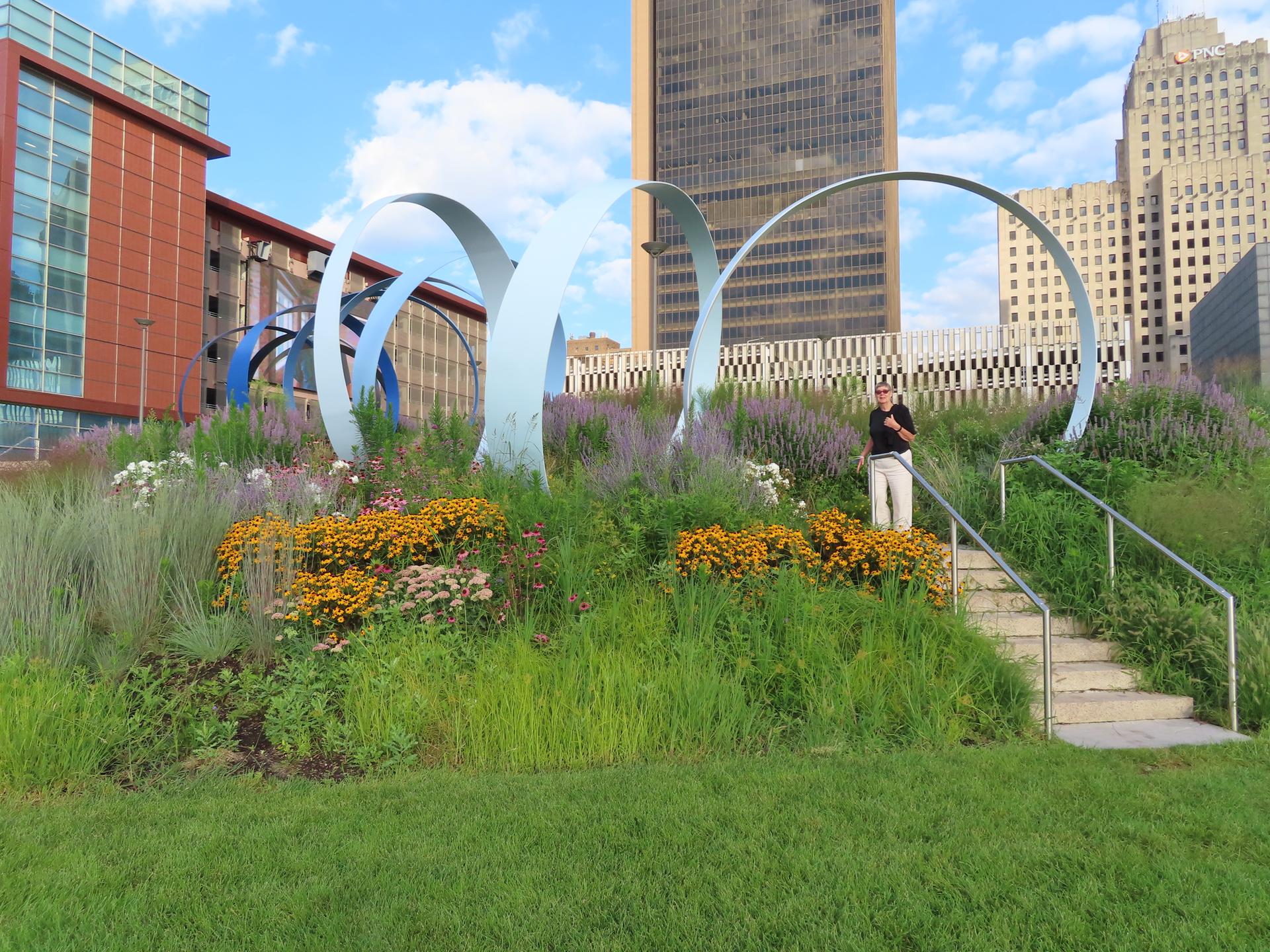Person stands by large white sculptures in a vibrant urban garden with buildings.