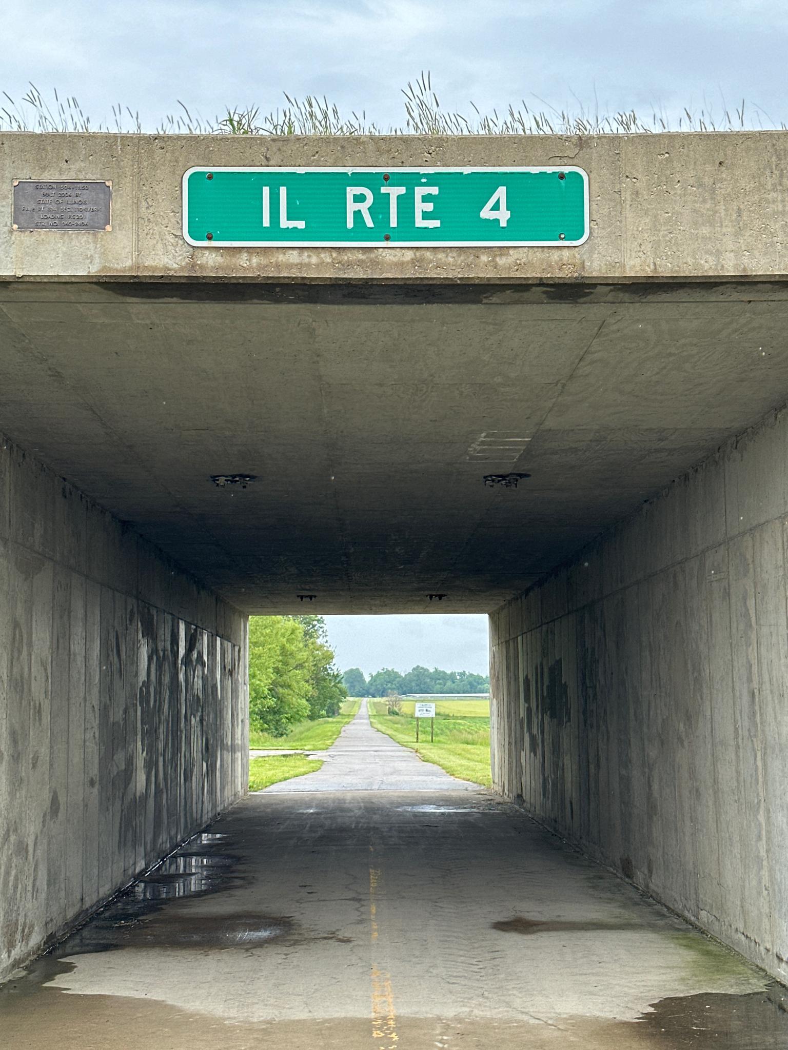 A paved path exits a concrete tunnel into a bright, open field landscape.