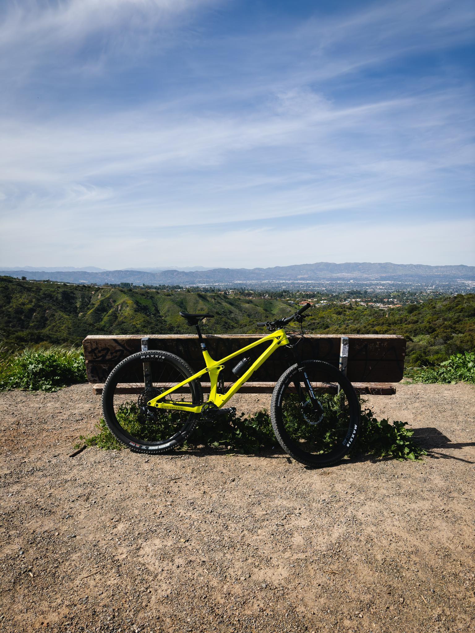 A bright yellow mountain bike rests on a bench overlooking green hills.