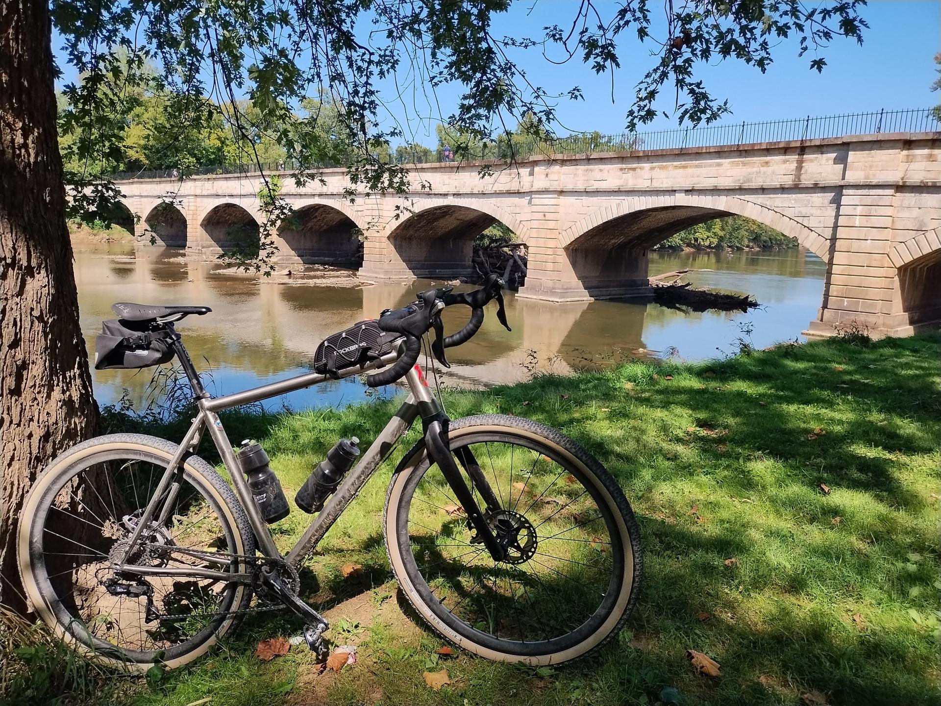 A bicycle leans against a tree with a stone arch bridge over a river.