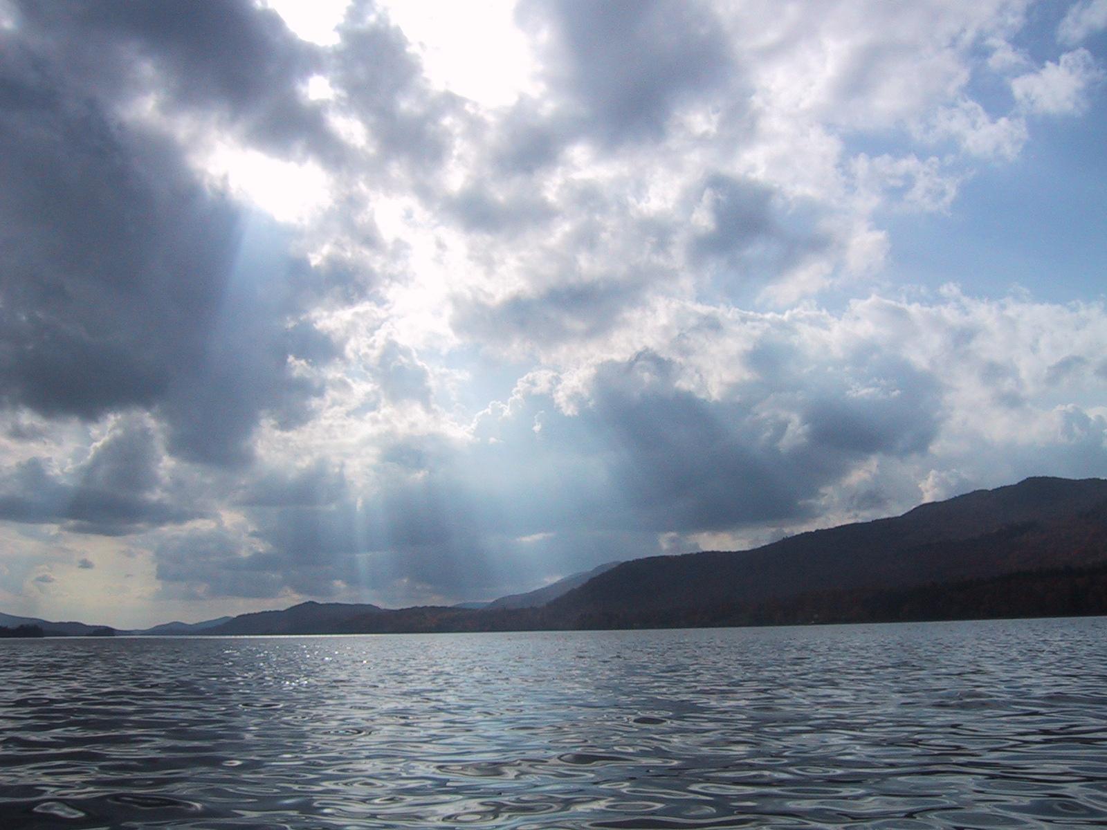 Sun rays pierce through dark clouds over a vast lake and distant mountains.