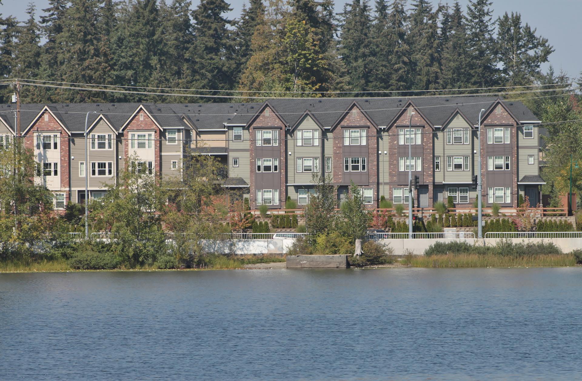 A row of modern townhouses overlooks a wide body of water and evergreen trees.
