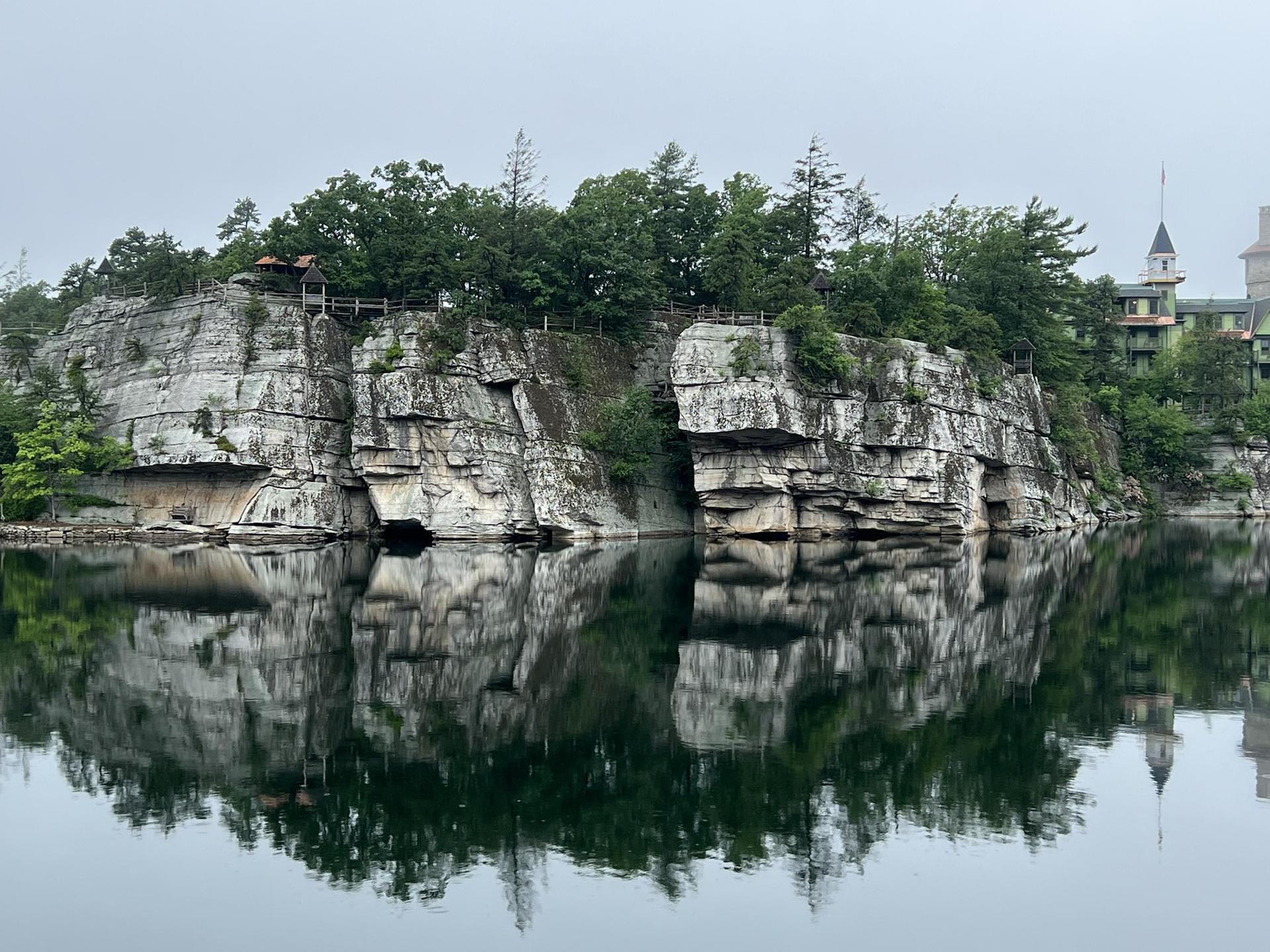 Calm lake reflecting rocky cliffs, green trees, and a building on an overcast day.