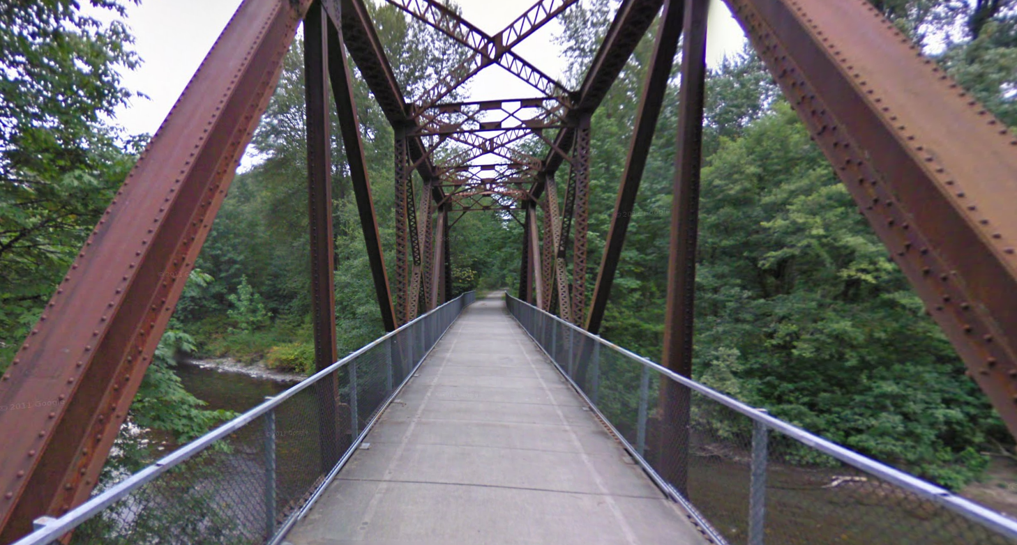 Rusty metal truss bridge with a path, surrounded by green trees. - Cedar River Trail