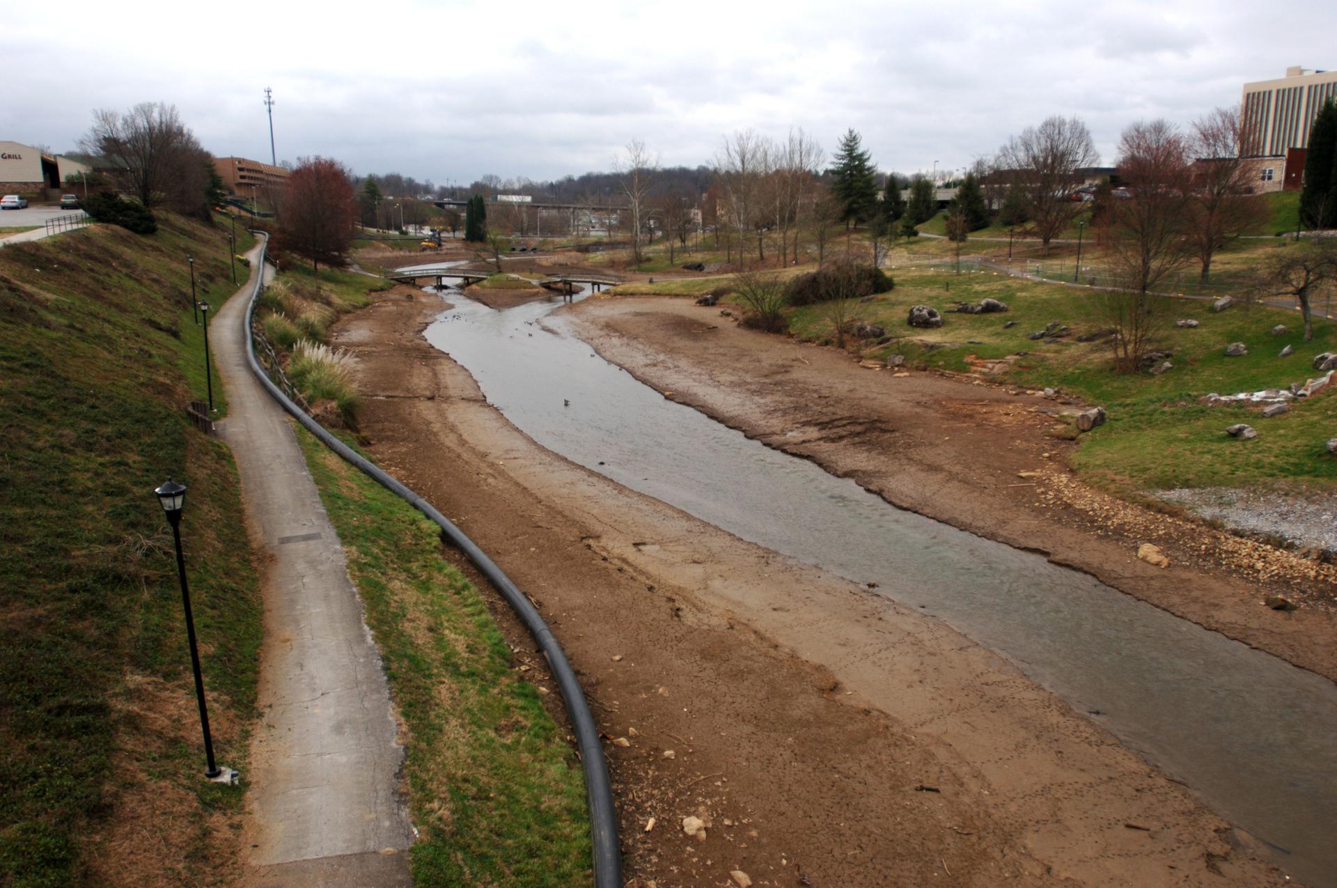 A narrow stream flows through a wide, mostly dry riverbed under an overcast sky.