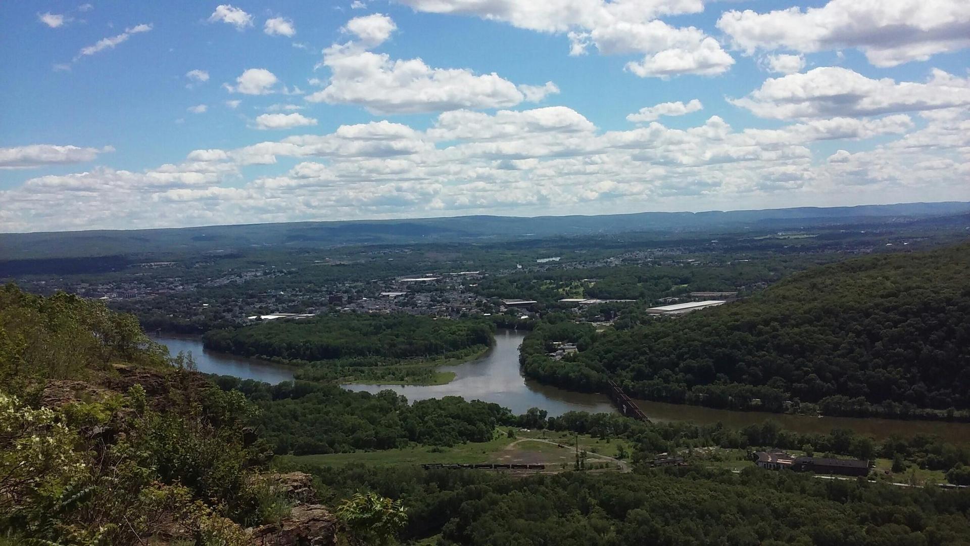 A panoramic view of a river, town, and hills under a cloudy sky.