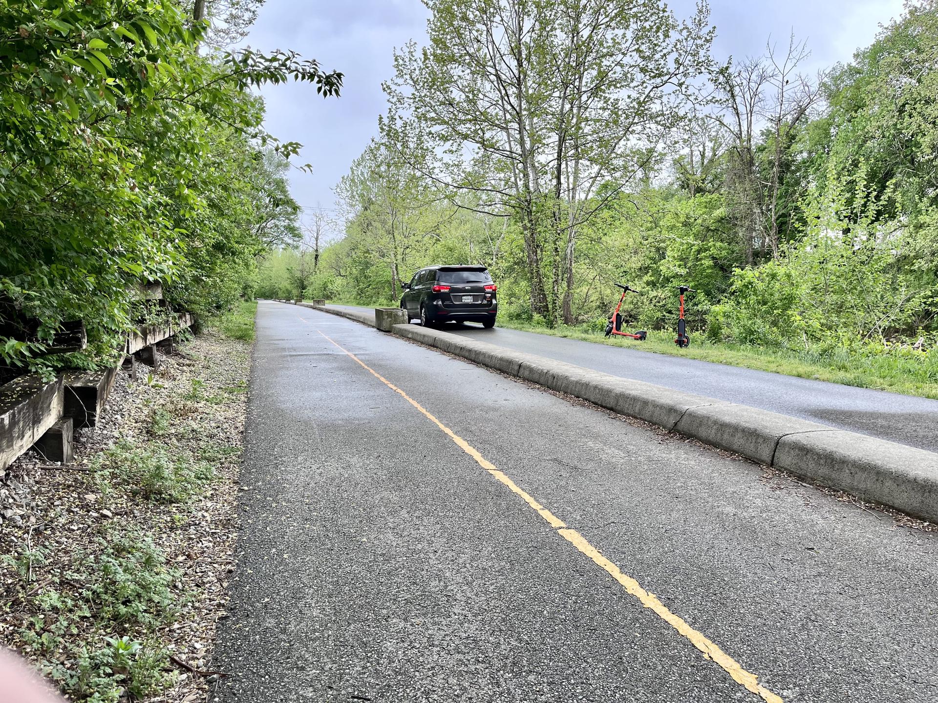 Paved path with a yellow line, parked car, and scooters under an overcast sky.
