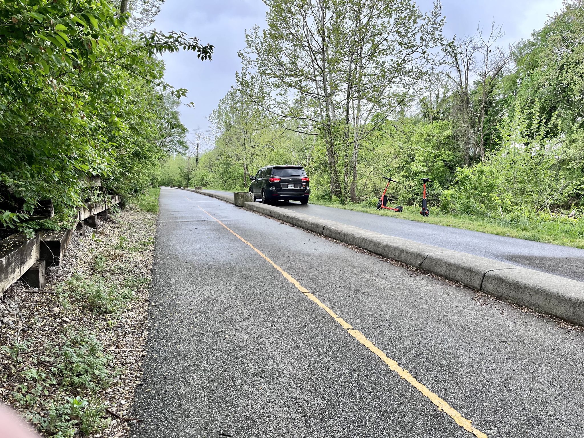 Paved path with a yellow line, parked car, and scooters under an overcast sky. - Roanoke River Greenway