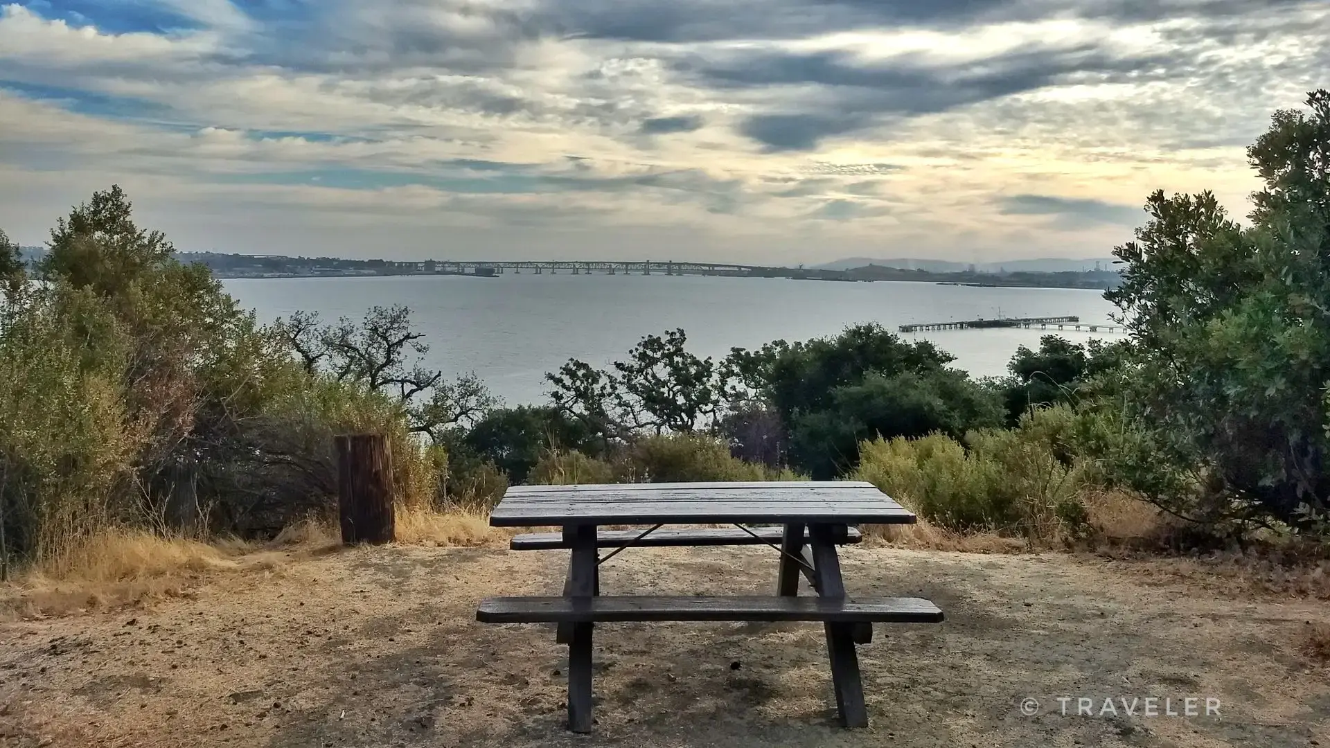 A wooden picnic table overlooks a wide bay under a cloudy sky.