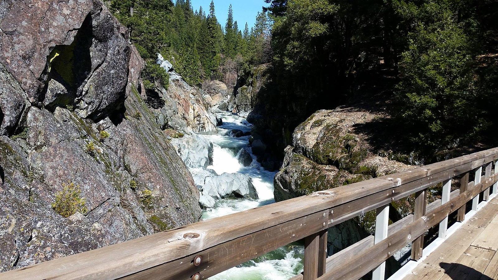Rushing river in a rocky gorge, viewed from a wooden bridge on a sunny day.