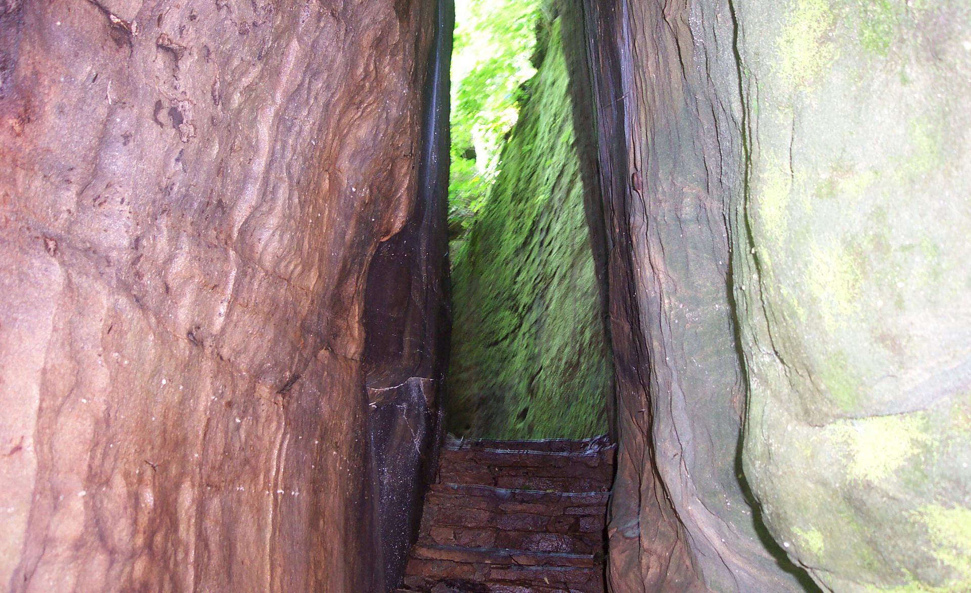 A narrow, dark rock crevice with wooden steps leading to bright green light.