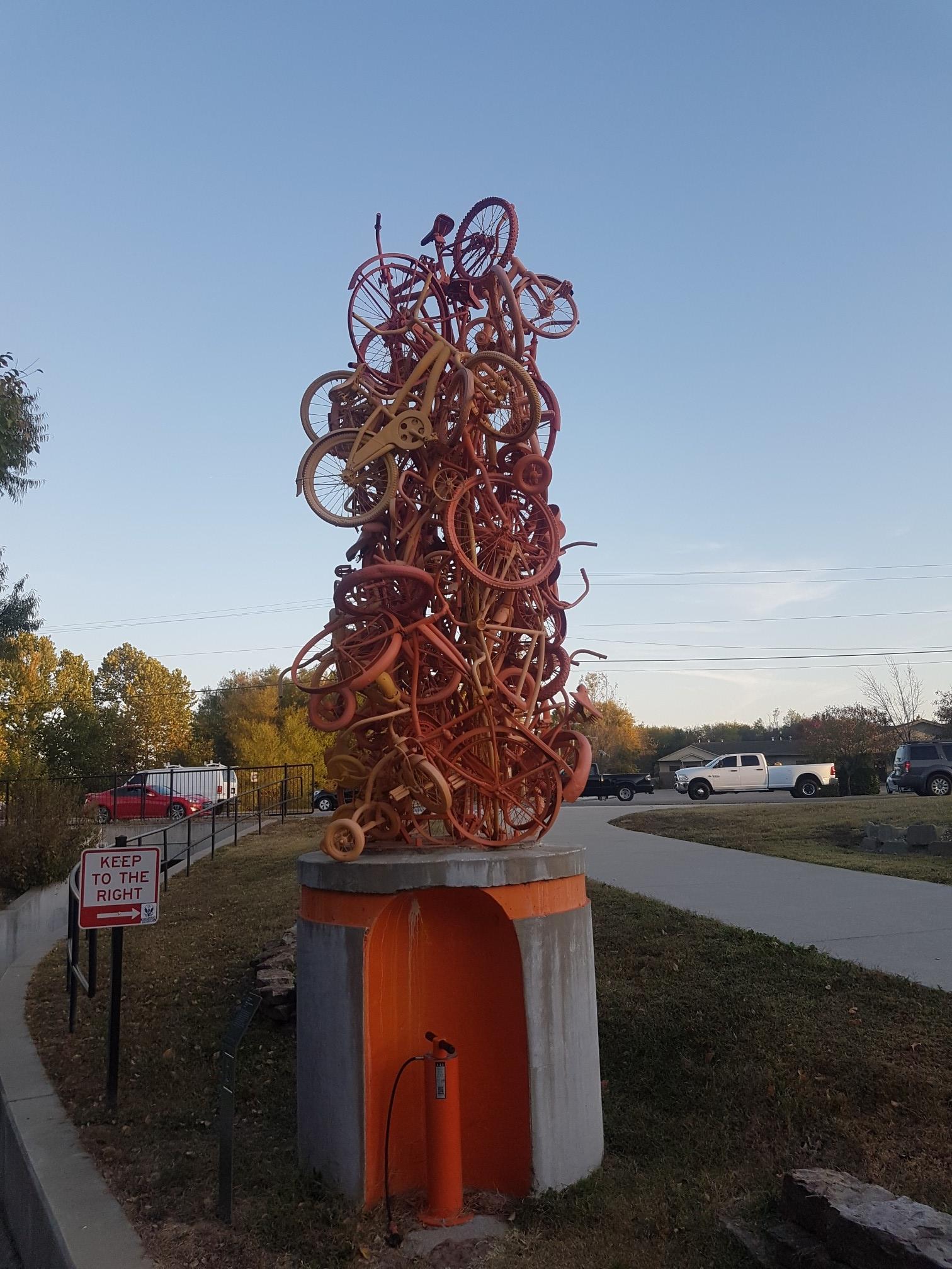 Tall sculpture of rusty bicycle wheels on a concrete base under a clear sky.