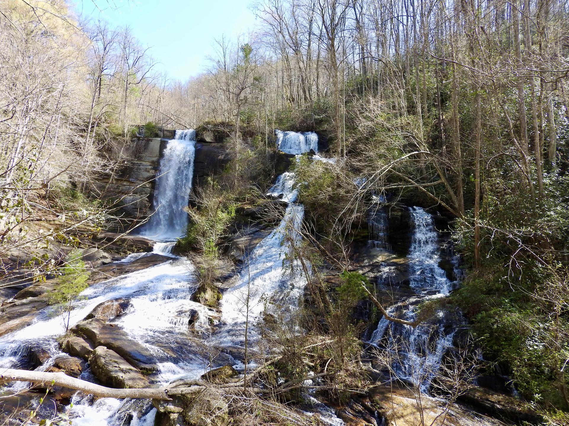 Multiple waterfalls cascade down a rocky, wooded hillside under bright daylight.