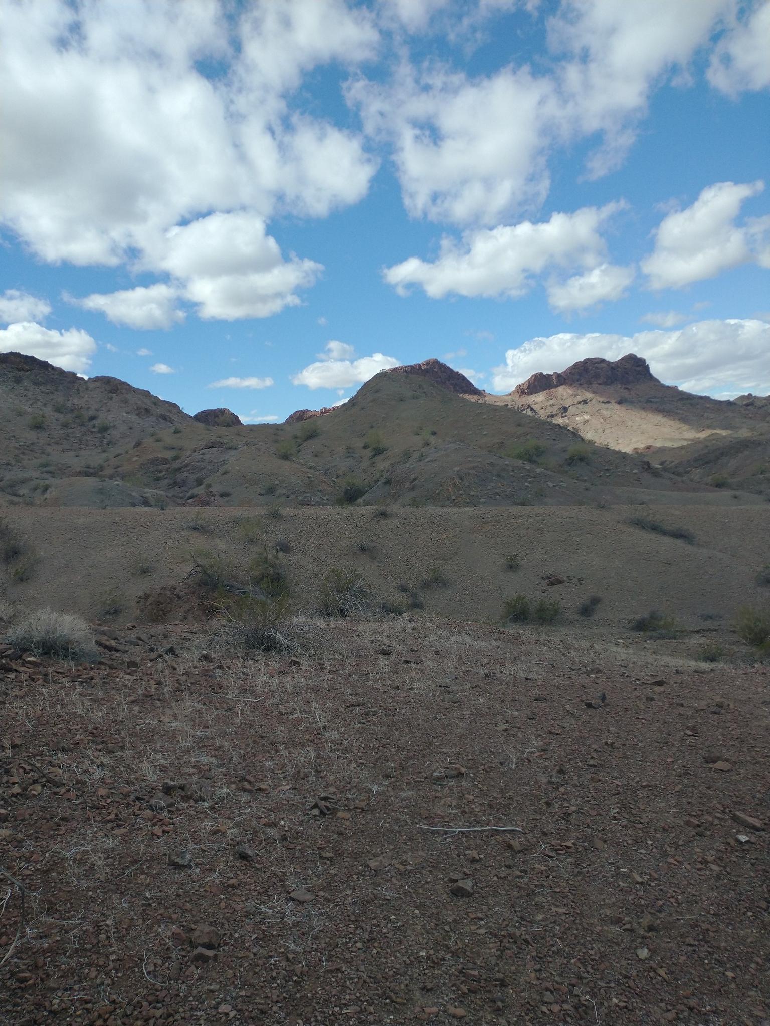 Desert hills with sparse vegetation and distant mountains under a bright cloudy sky.