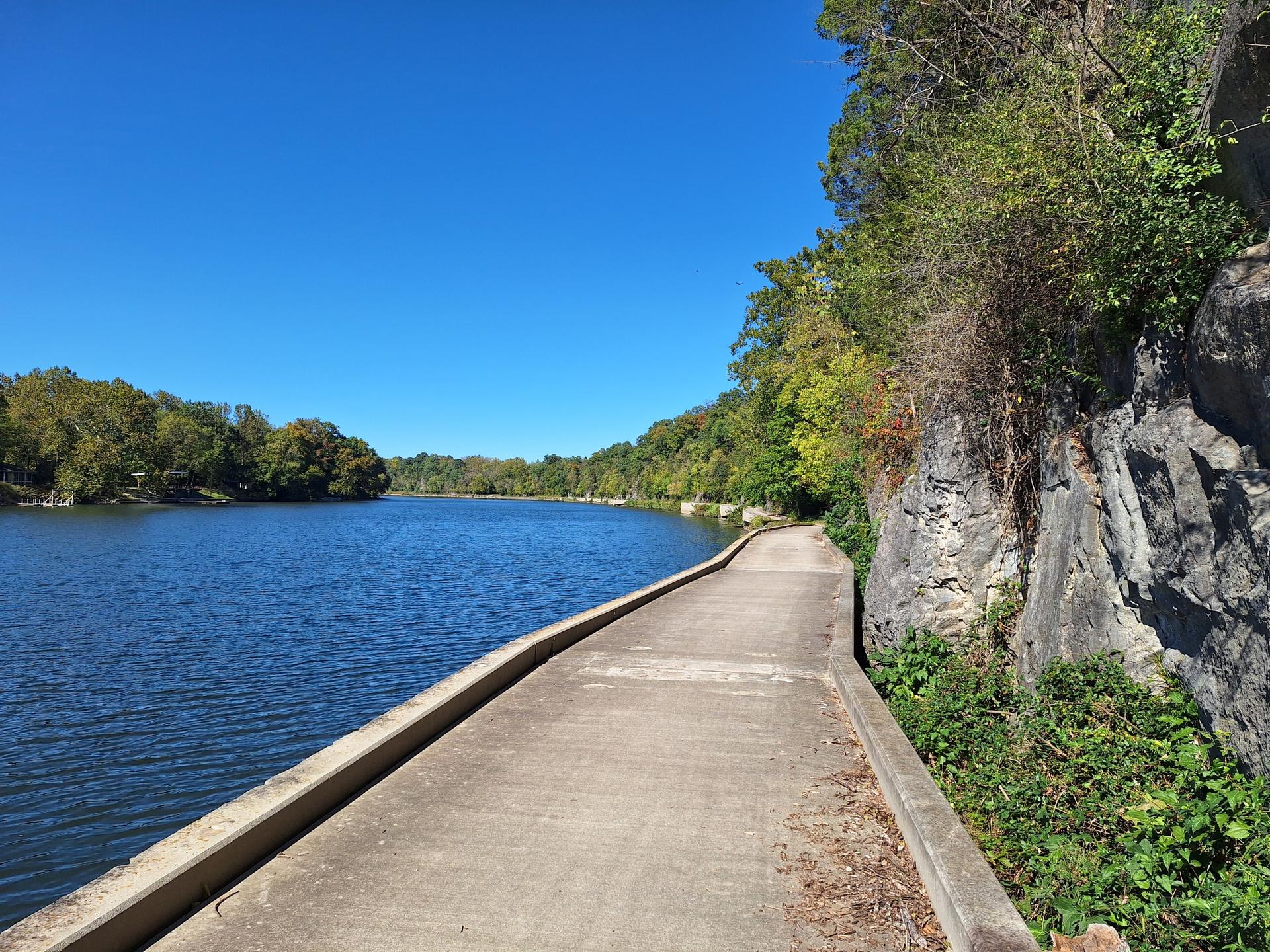 Concrete path next to a rock wall and a wide, calm river.