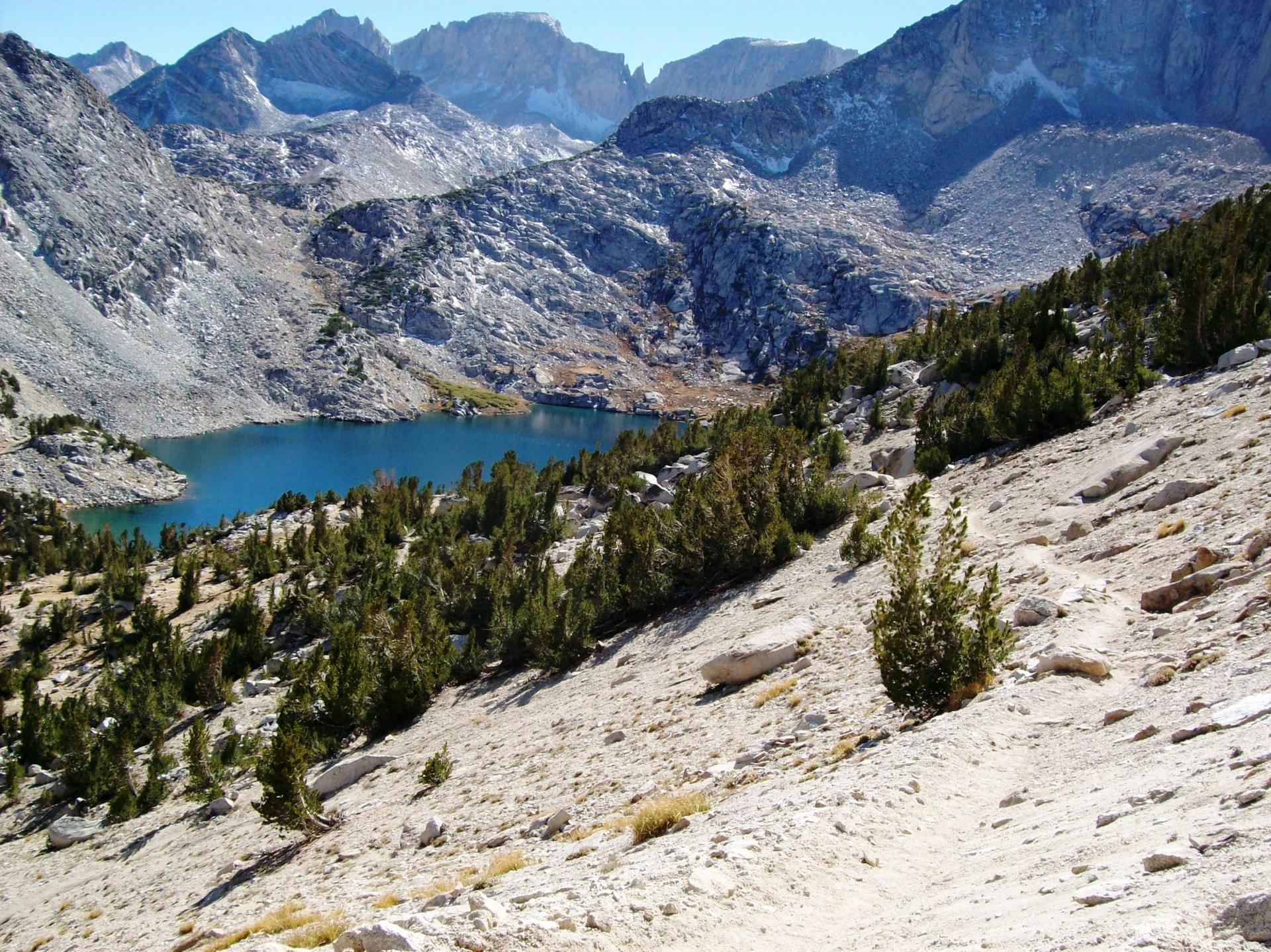 Steep rocky hillside descends to a blue lake surrounded by mountains.