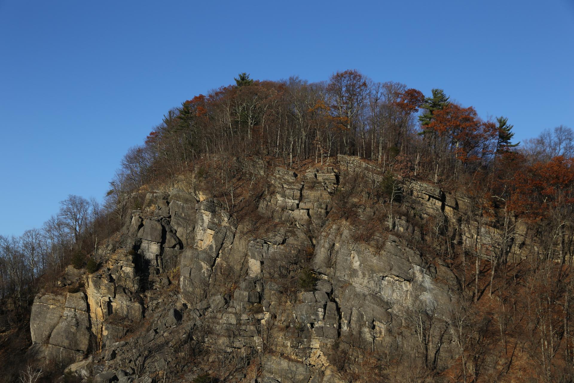 Steep rocky cliff with bare trees and some autumn foliage under a clear sky.