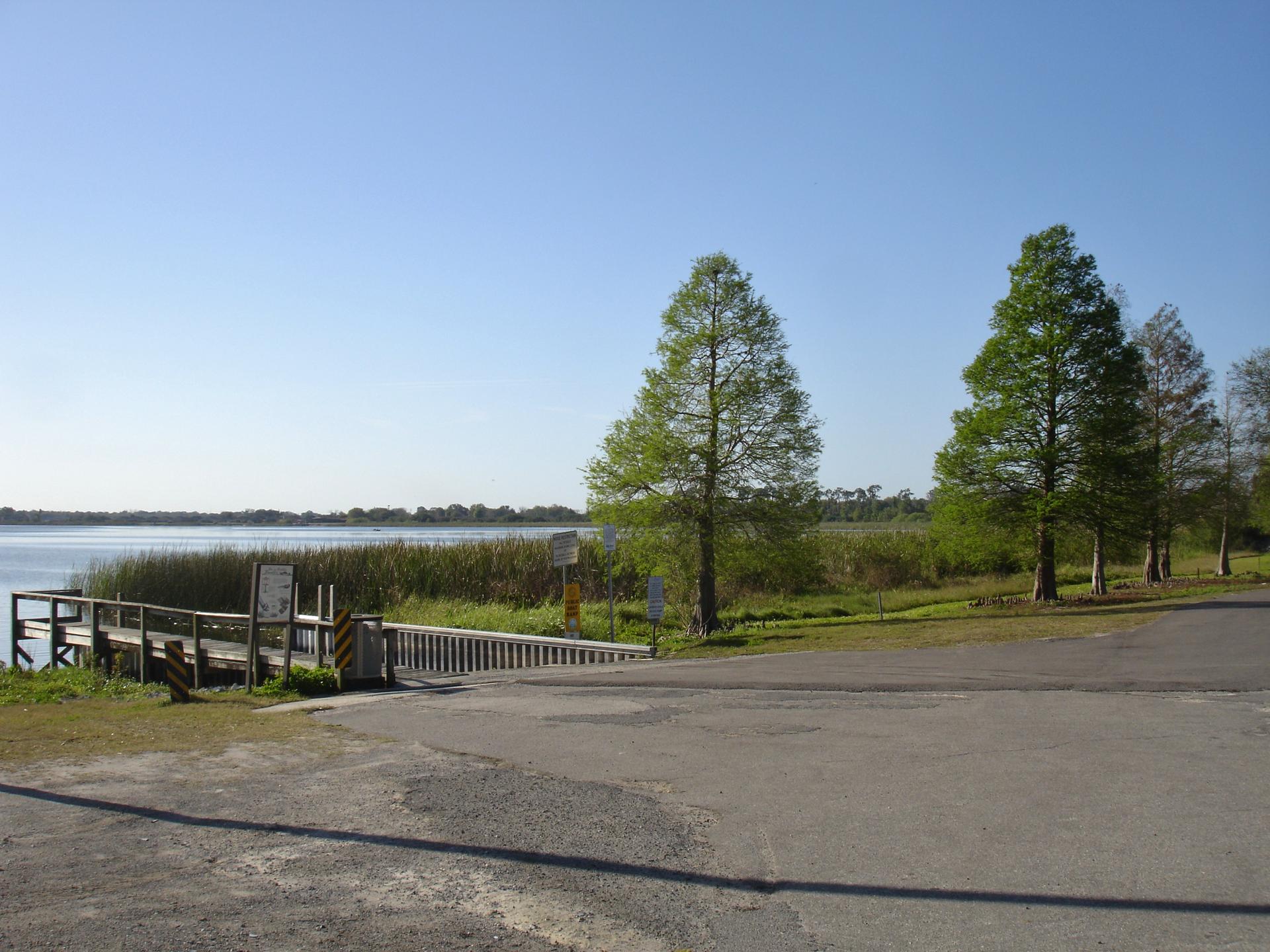 A boat ramp and wooden dock by a lake with tall green trees.