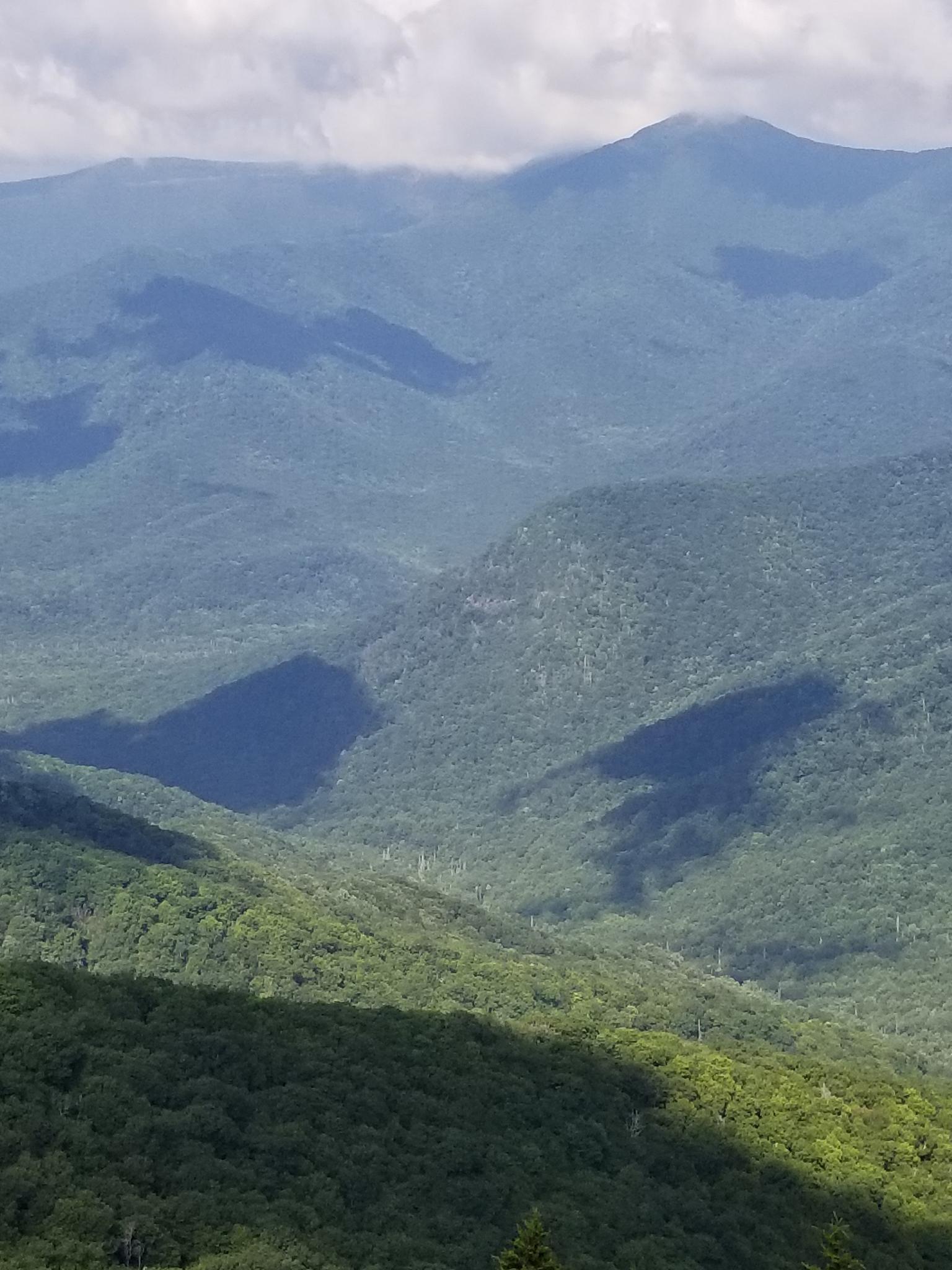 Vast forested mountains and valleys under a partly cloudy sky with dappled shadows.