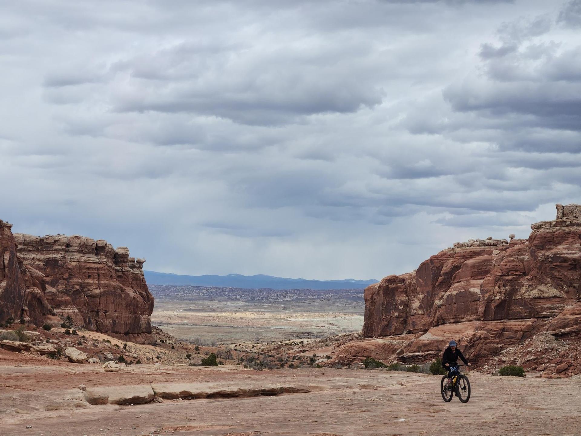 A person mountain biking through a wide desert valley under an overcast sky.