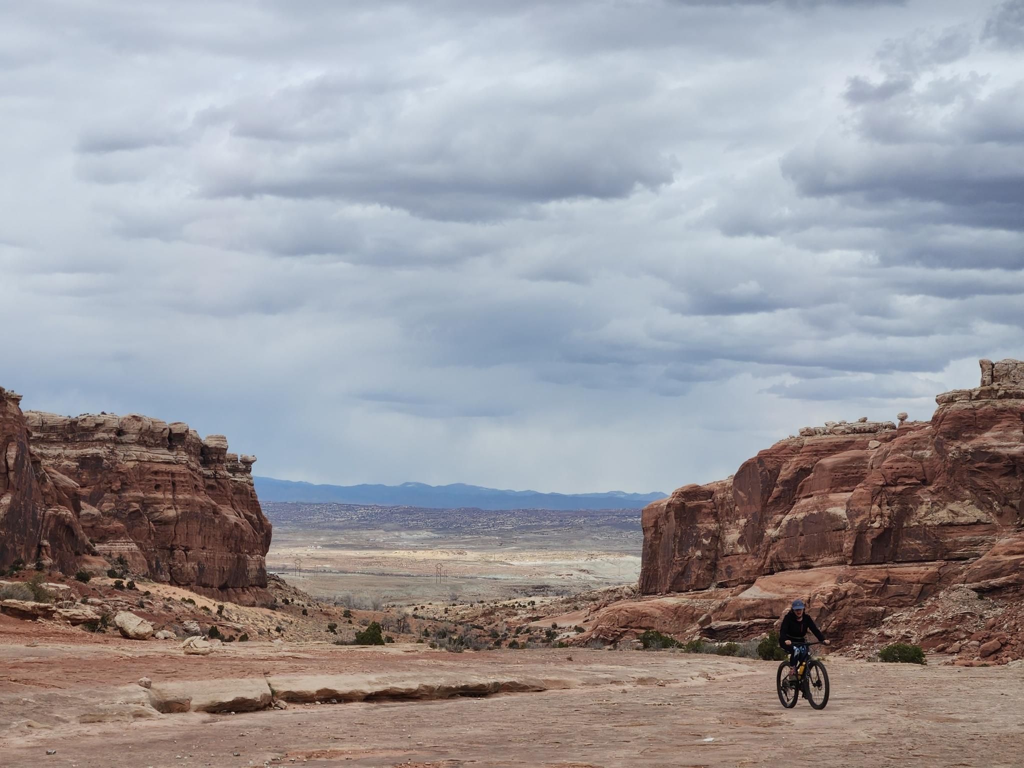 A person mountain biking through a wide desert valley under an overcast sky. - Lower Monitor & Merrimac Trail Loop